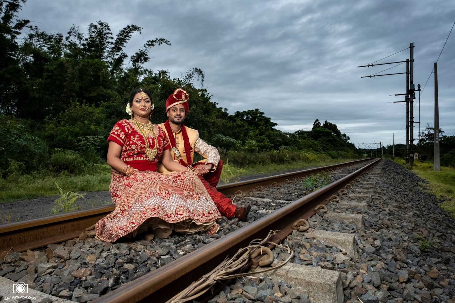 Traditional Indian wedding couple in red attire pose on railroad tracks under moody skies.