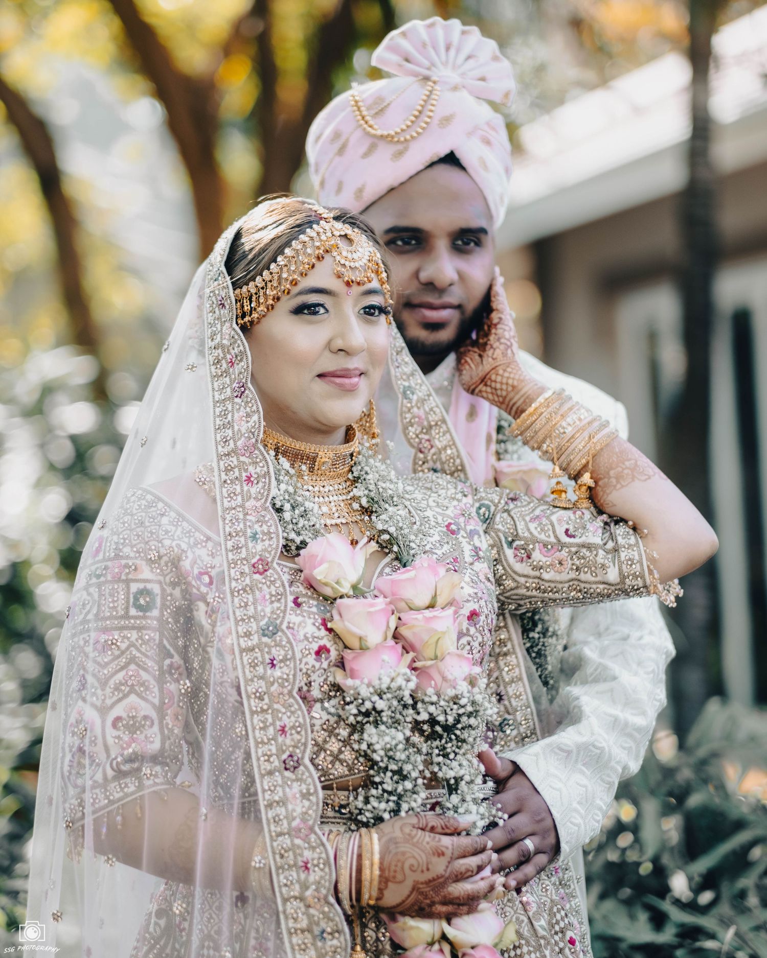 Couple in white traditional wedding attire posing with pink floral bouquet outdoors.