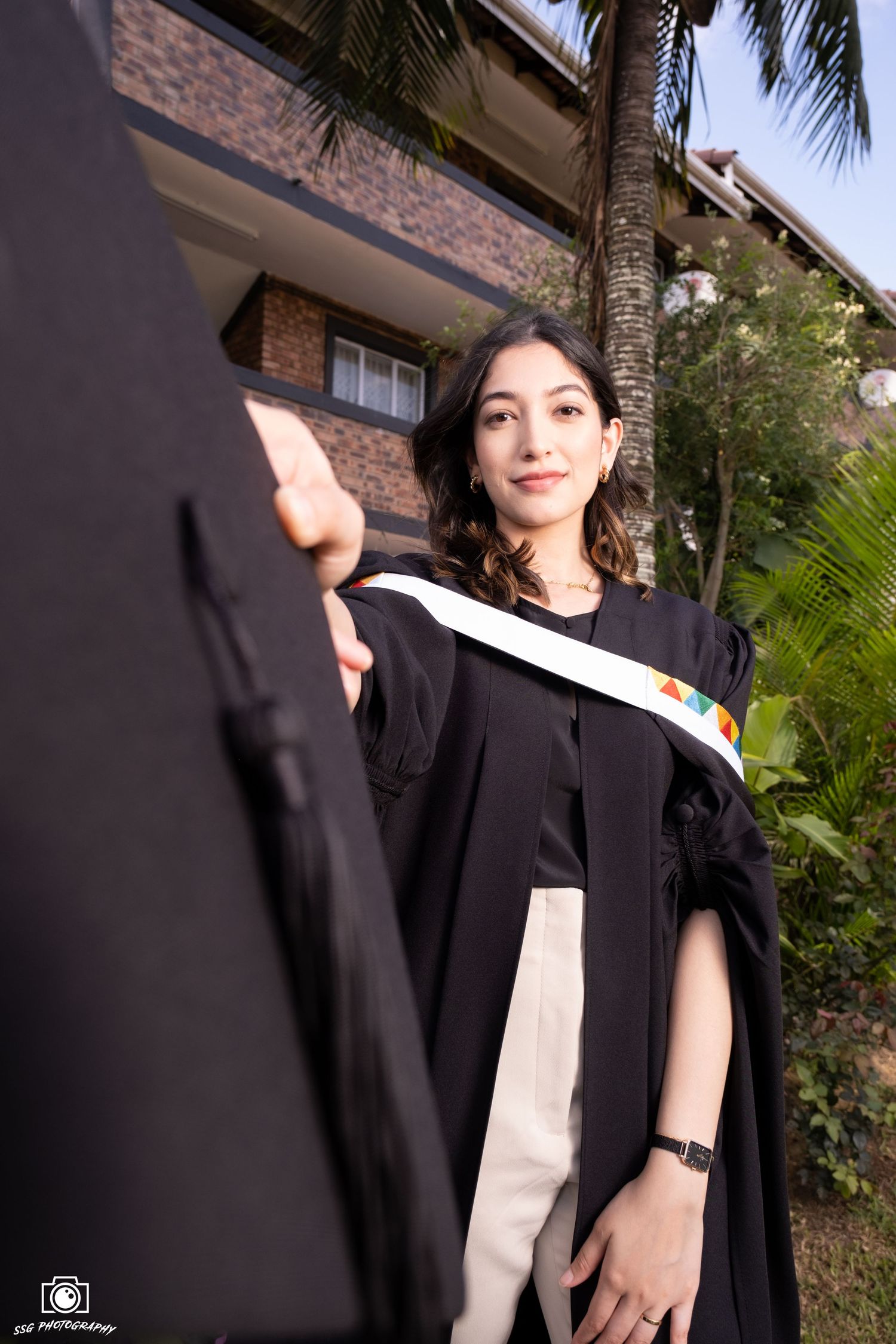 Graduate in black academic regalia poses outdoors near palm trees and brick wall.