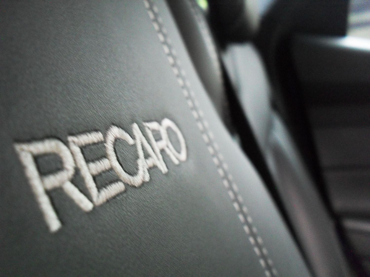 Close-up detail of Recaro logo embroidered in white stitching on gray leather car seat upholstery.
