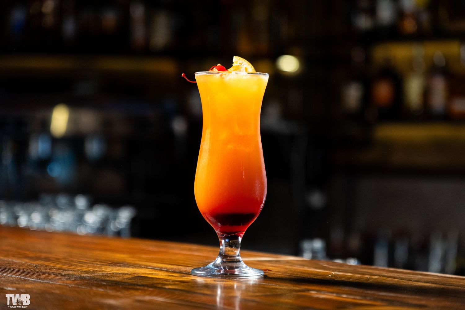 Layered orange and red tropical cocktail in a hurricane glass on dark wooden bar.