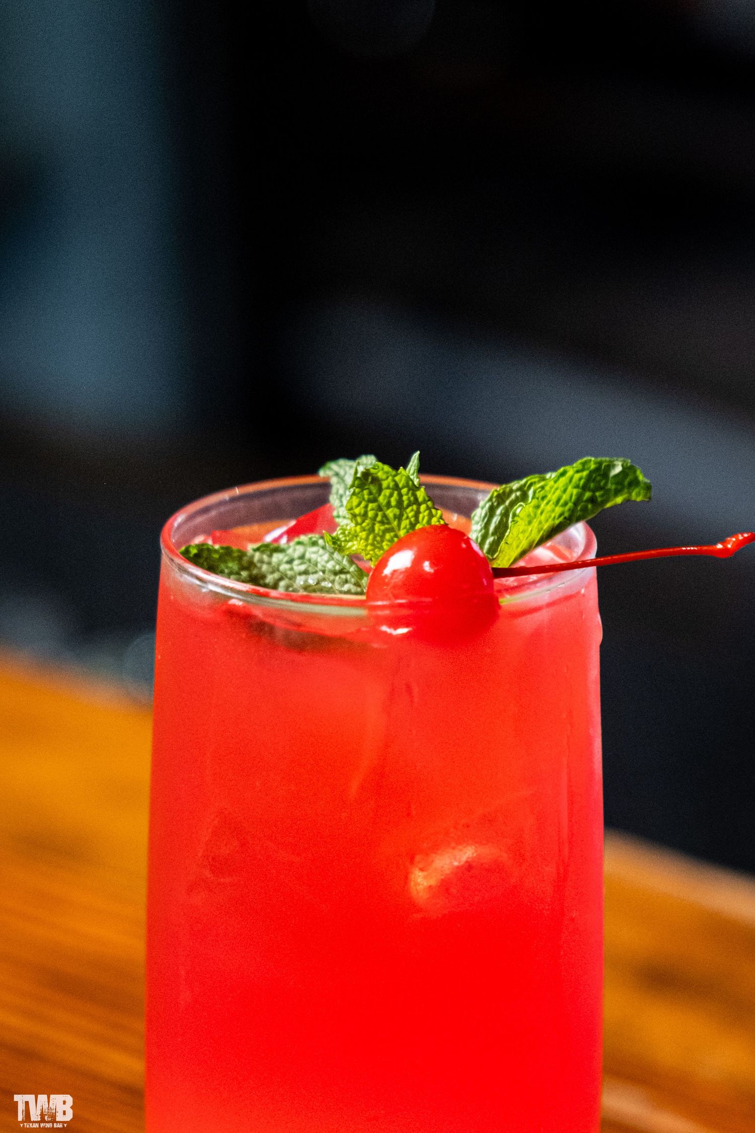 Bright red cocktail garnished with mint leaves and cherry on wooden bar surface.