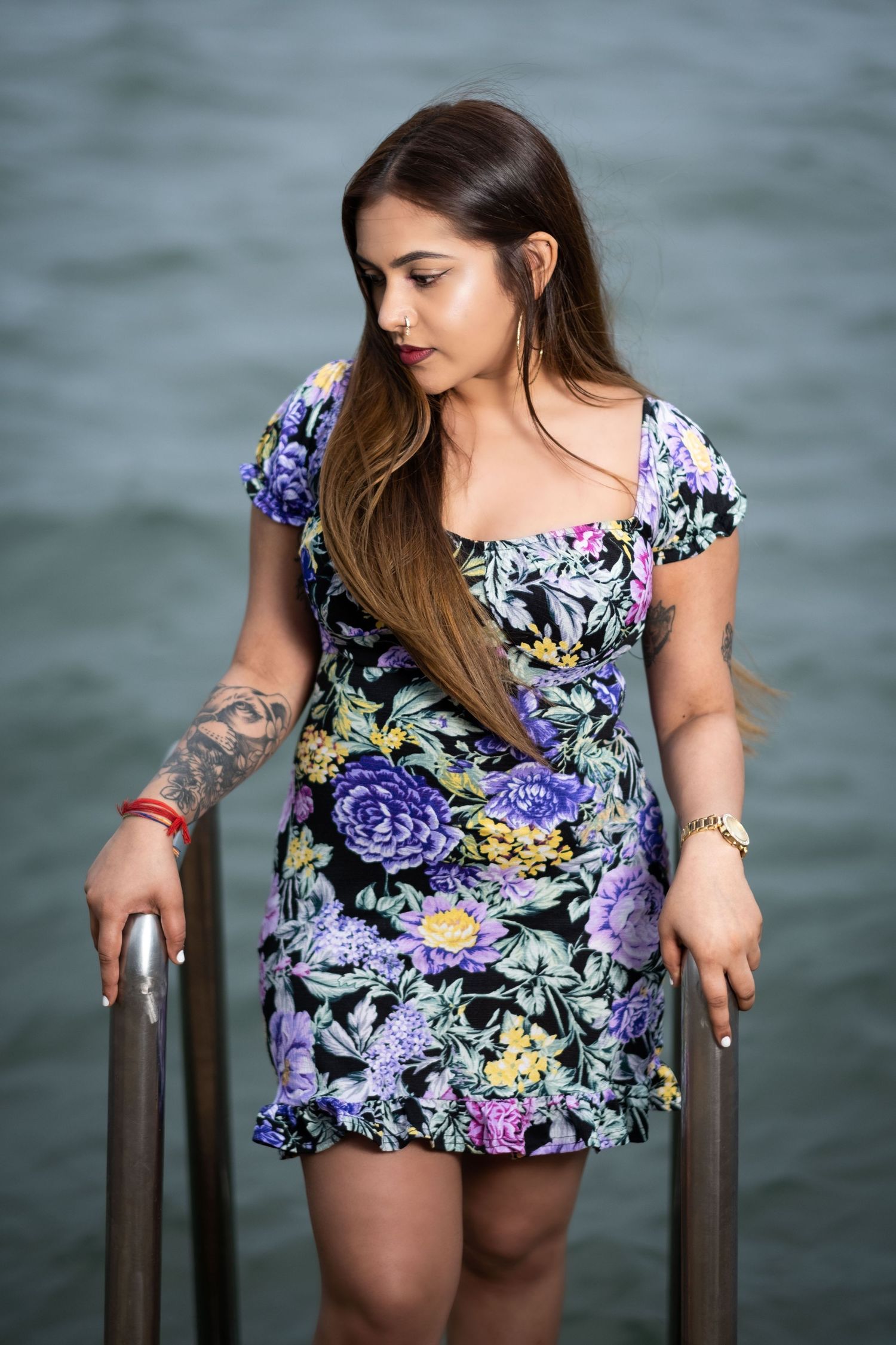Woman in purple floral dress poses gracefully on metal railing by the sea.