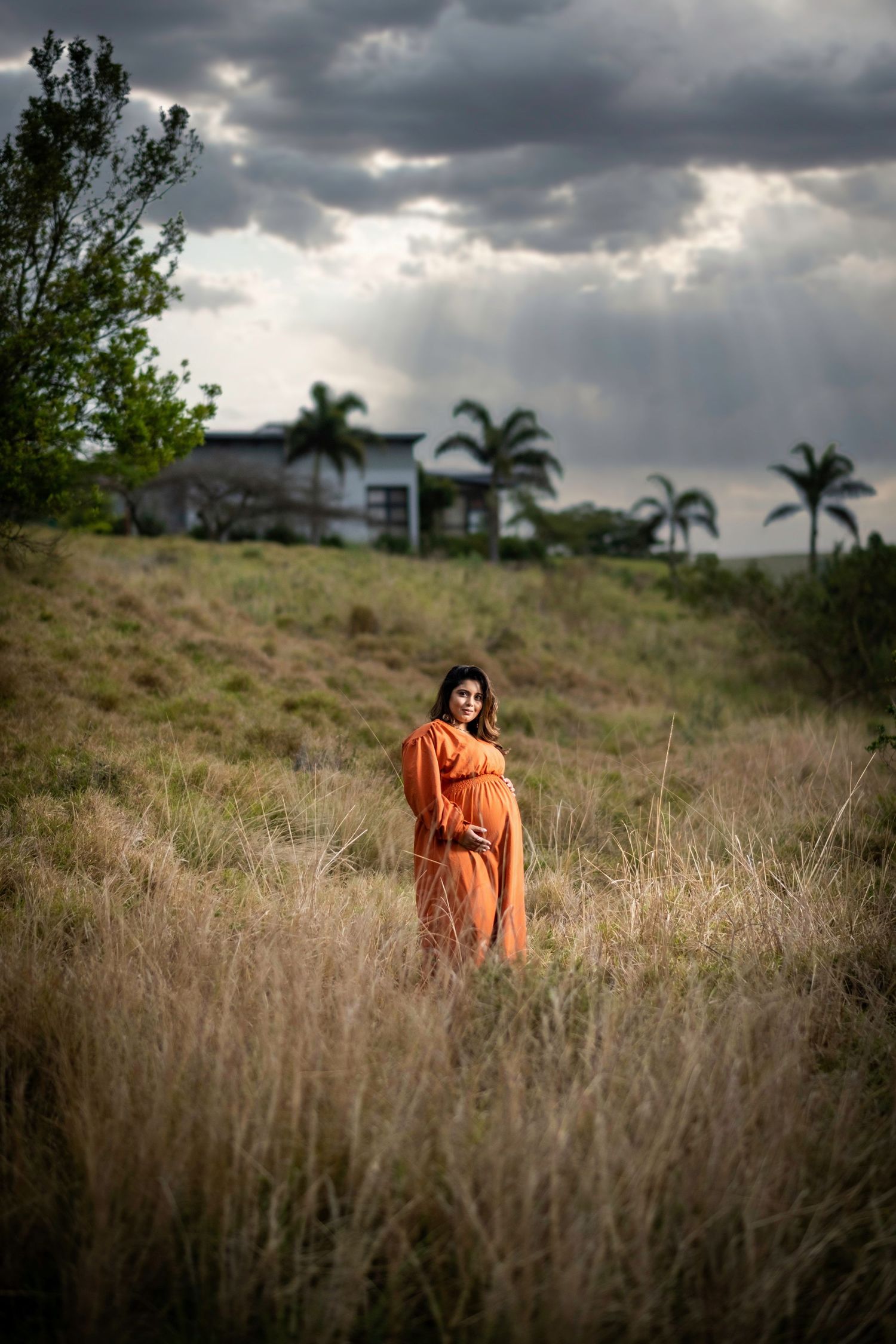 A figure in a flowing orange dress stands in tall grass with palm trees and dramatic cloudy skies in the background.