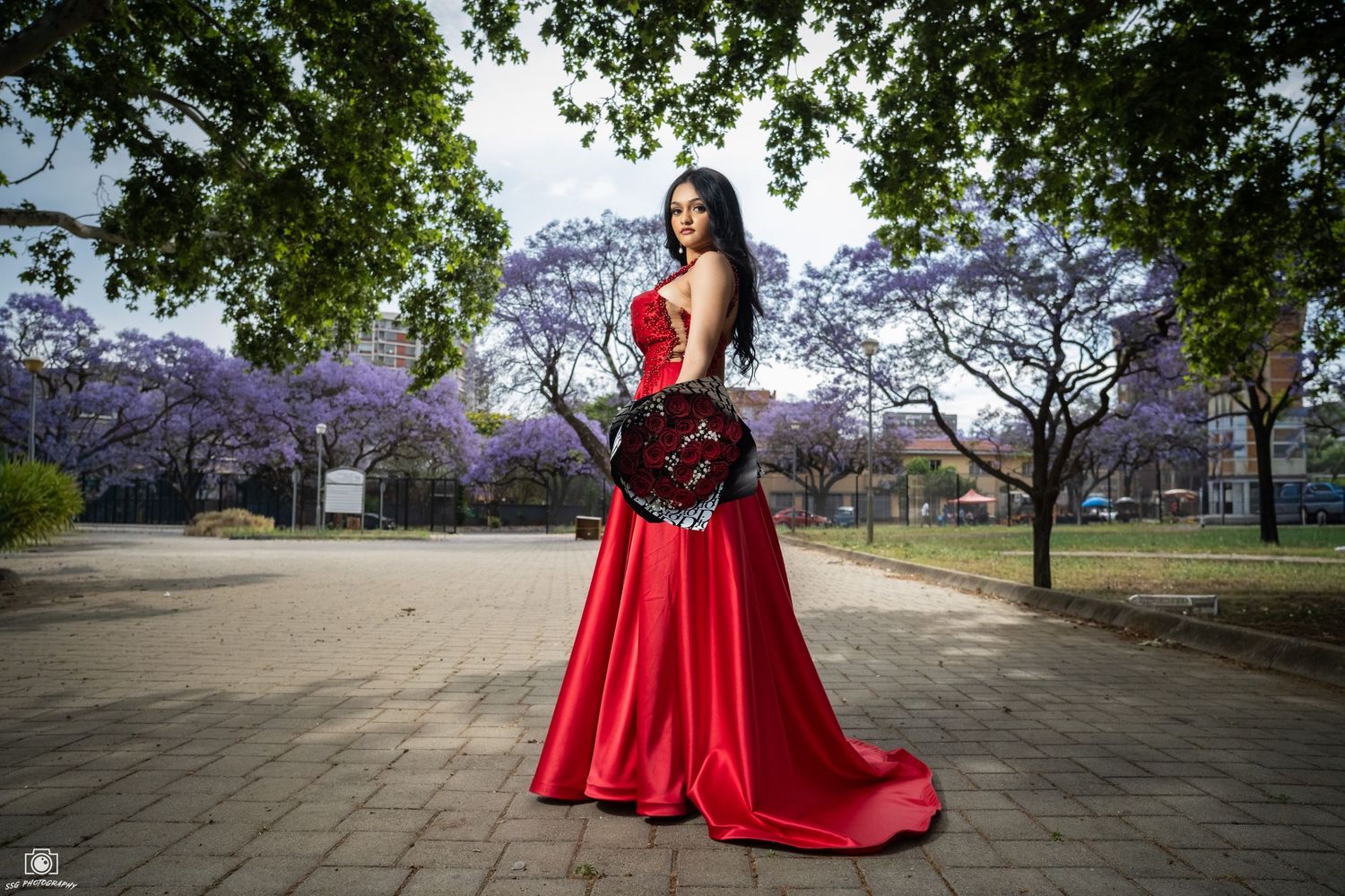 Outdoor fashion photography with a flowing red gown in a park setting under trees.