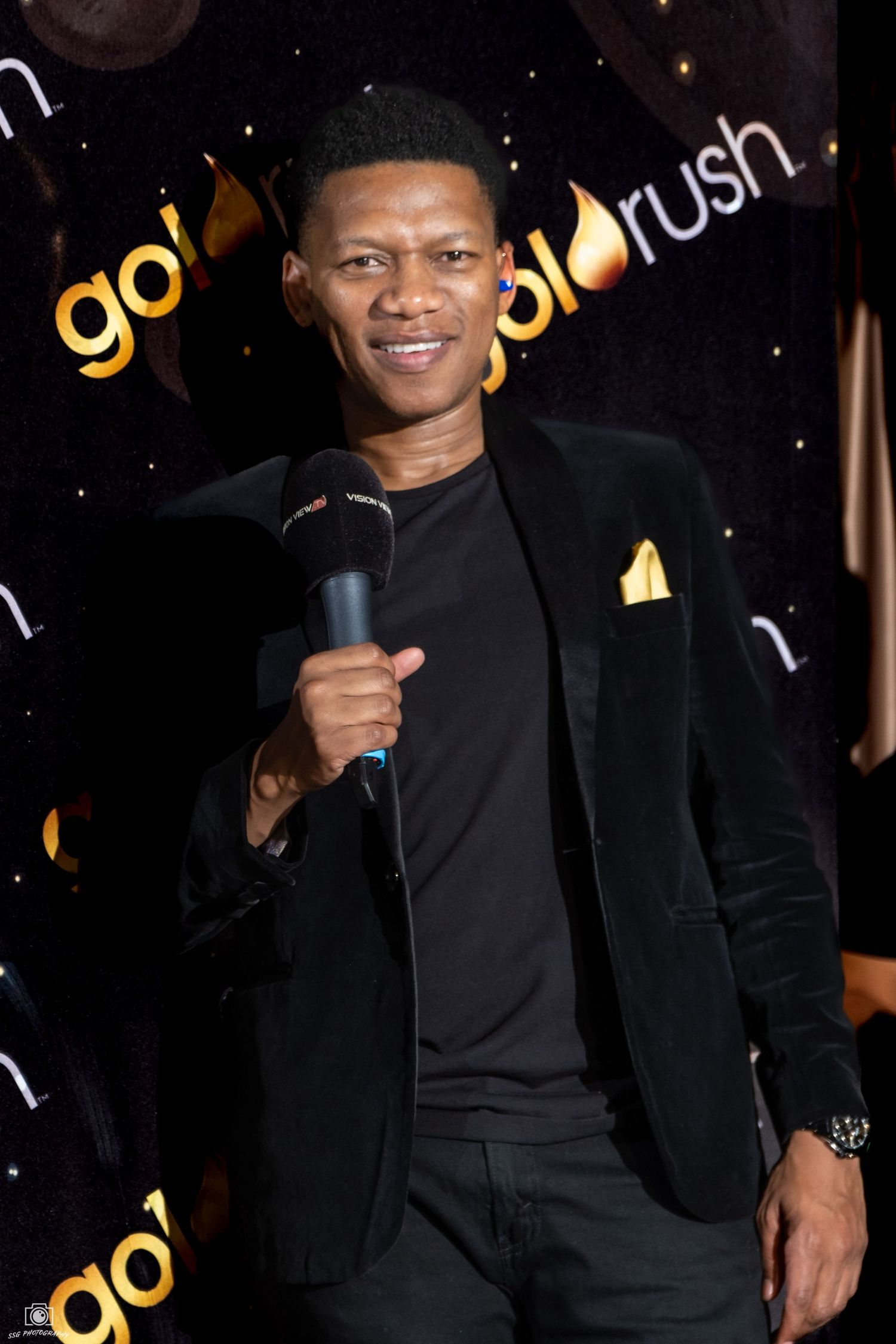 Performer in black blazer smiles while holding microphone against Goldrush branded backdrop.