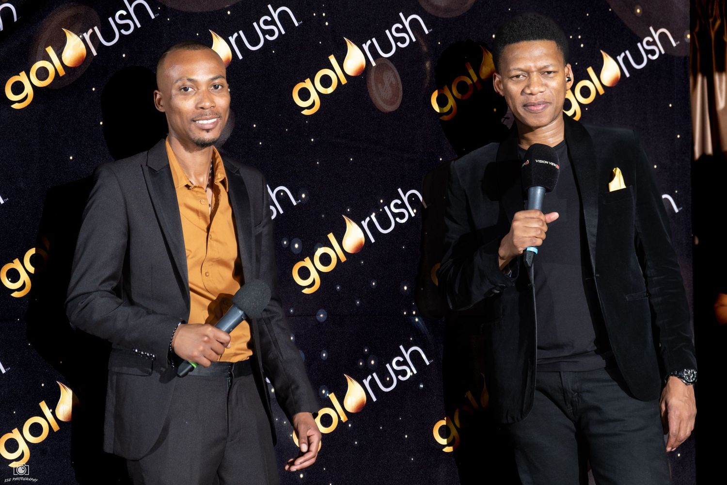 Two performers speak on stage at a Goldrush event with branded backdrop.