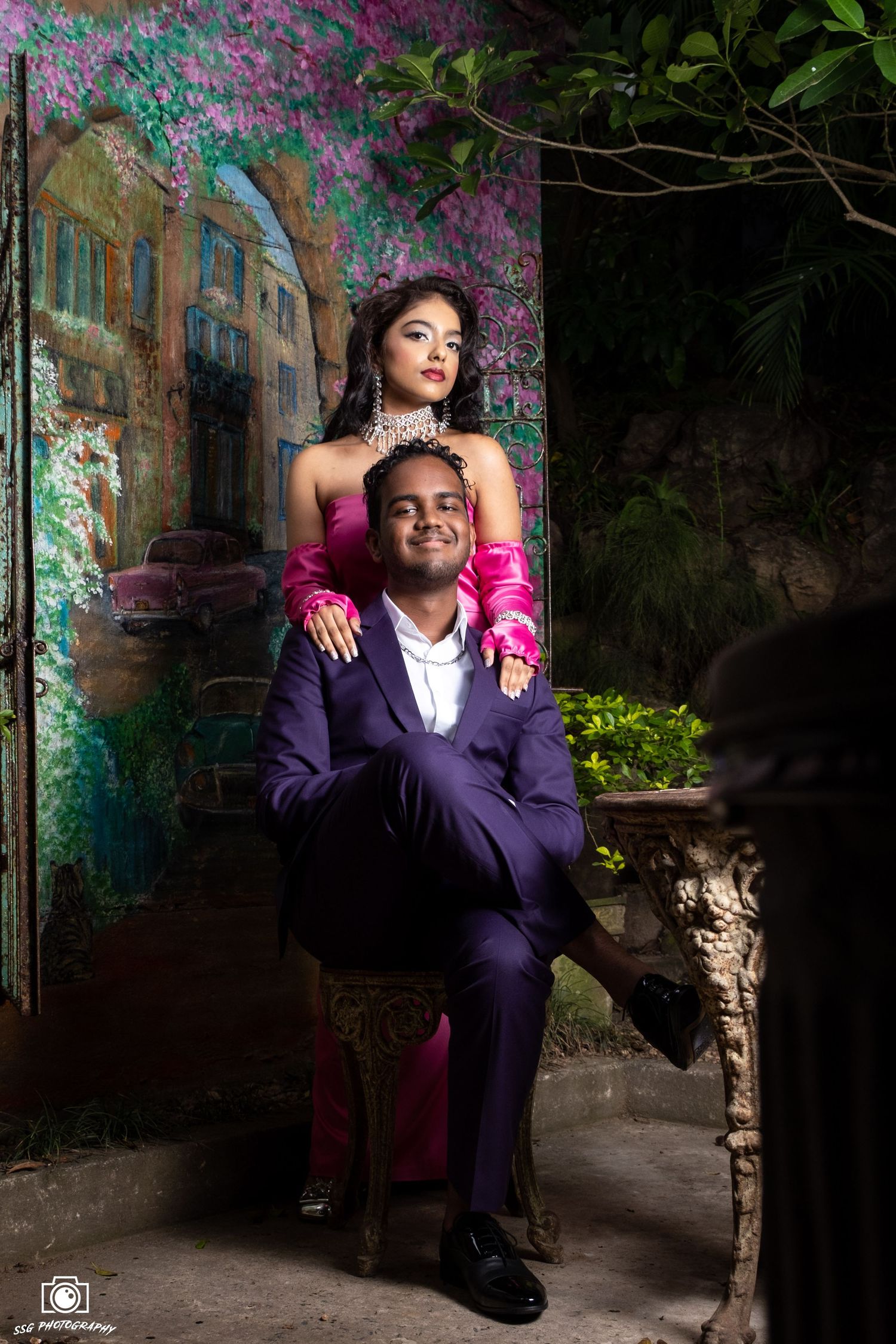 Couple in formal attire pose together against a colorful garden backdrop with purple flowers.