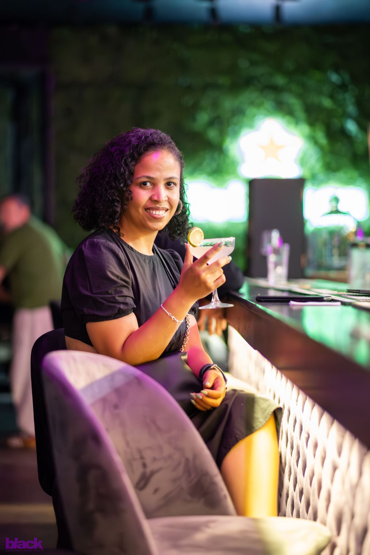 Woman enjoys cocktail while sitting at illuminated bar counter in evening setting.
