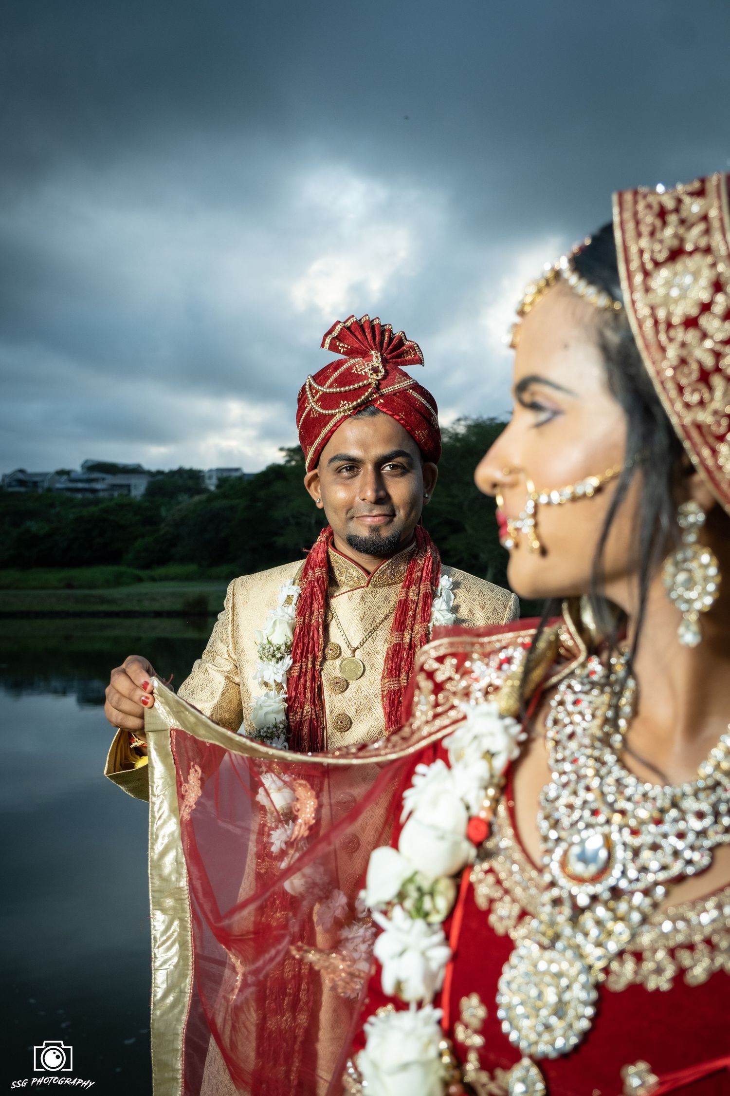 Traditional Indian wedding couple in ornate red and gold attire against dramatic stormy sky.