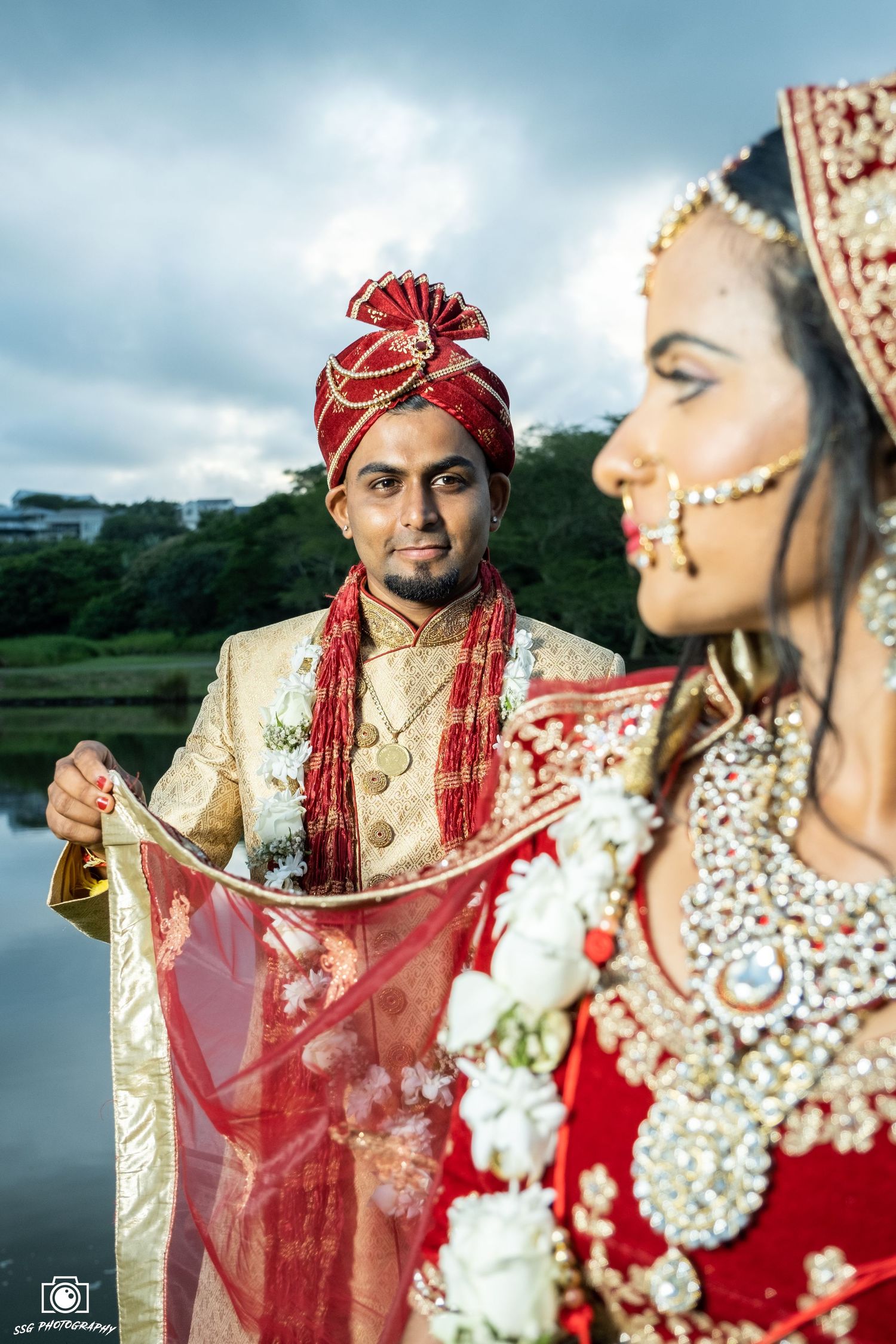 Traditional Indian wedding couple posing outdoors with red dupatta flowing in breeze.