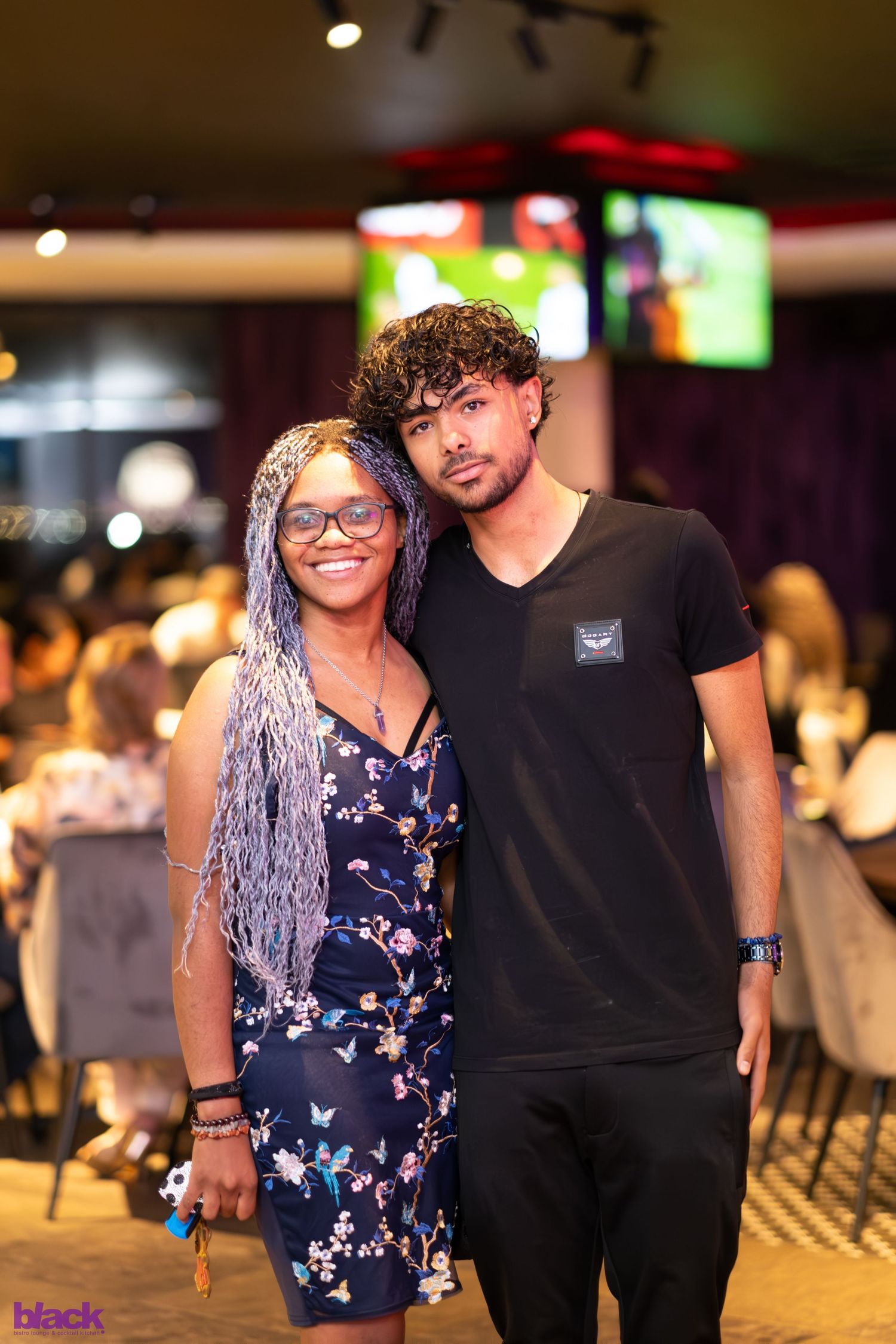 Two people pose together at a casual indoor event with TV screens visible in background.