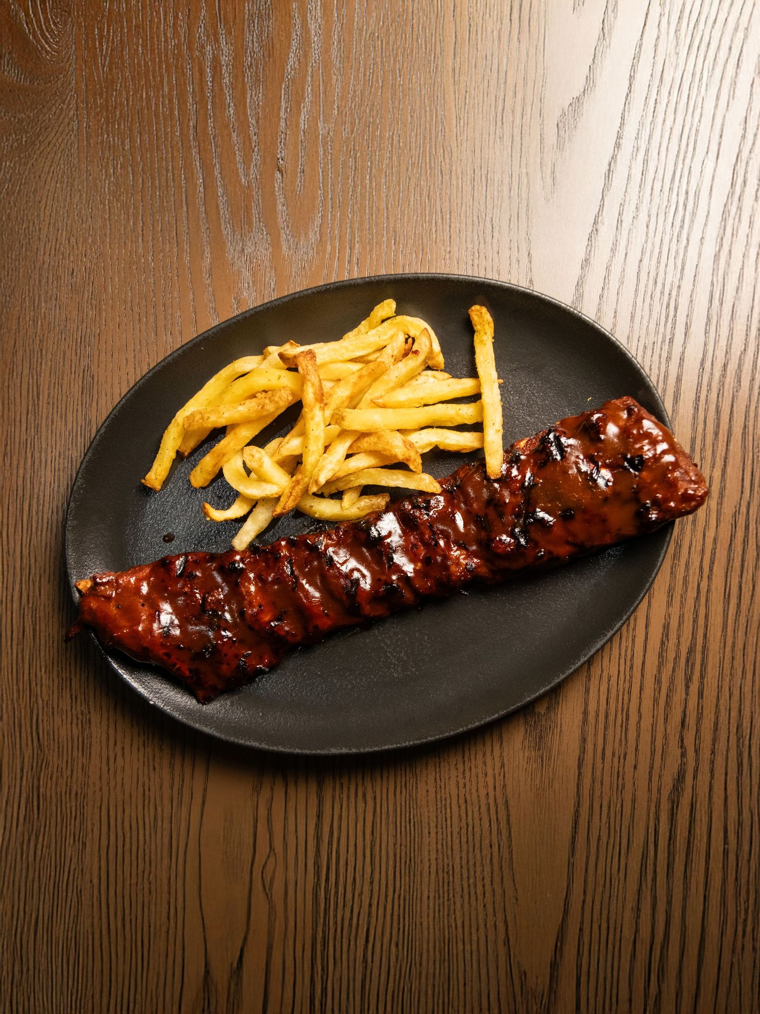 Glazed BBQ ribs with french fries served on black oval plates.
