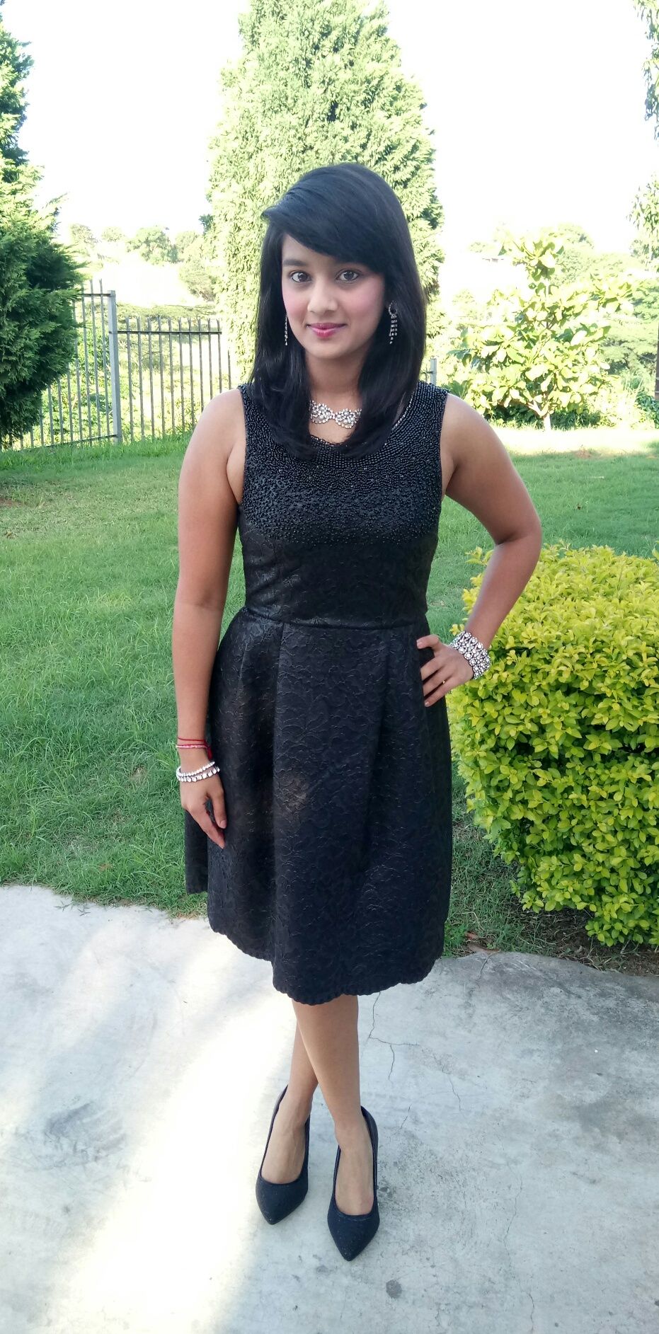 A person in a sleeveless black dress and heels poses outdoors on a paved area near green shrubbery.