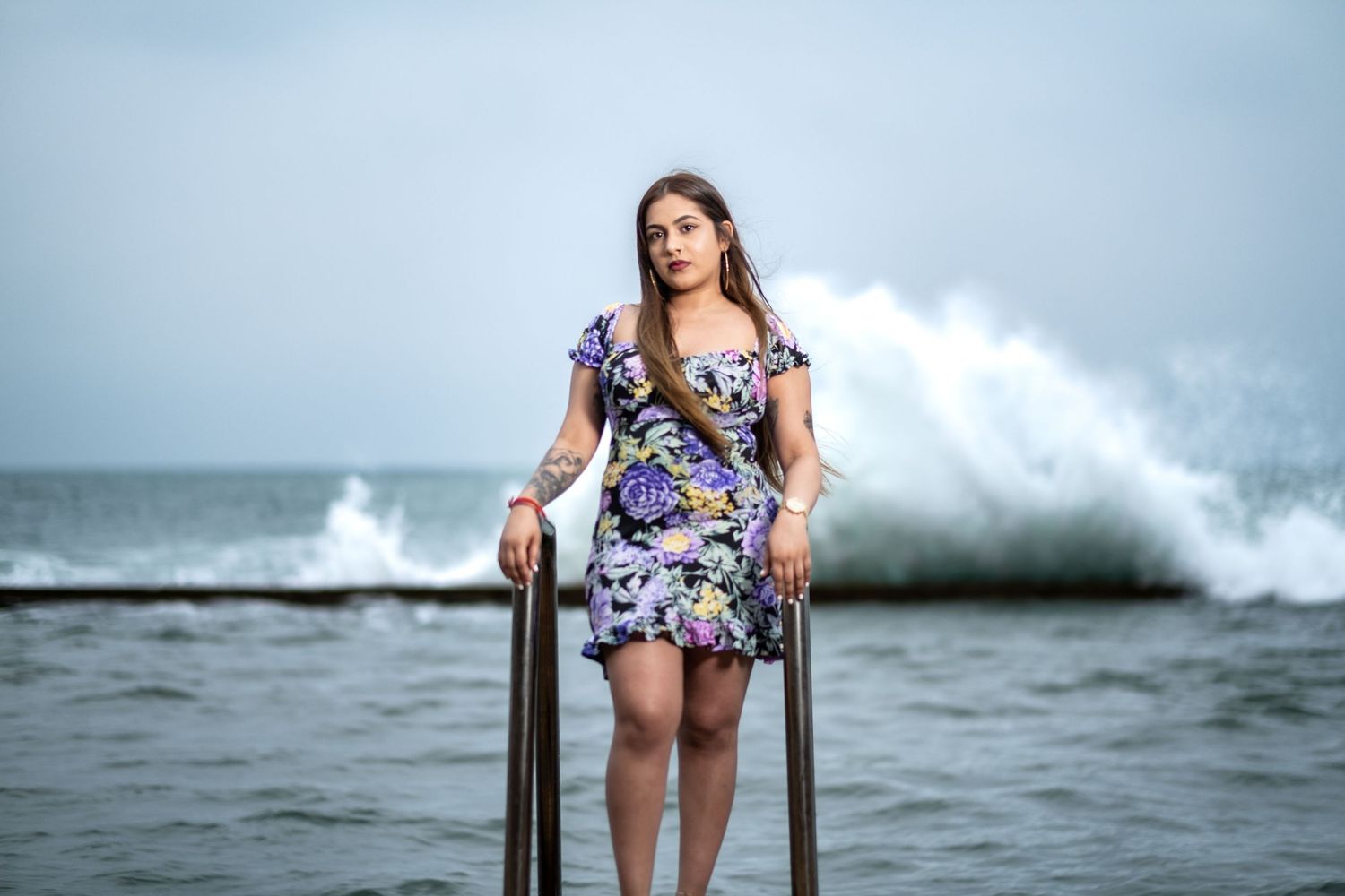 Model in floral dress sits on railing with crashing ocean waves in background.