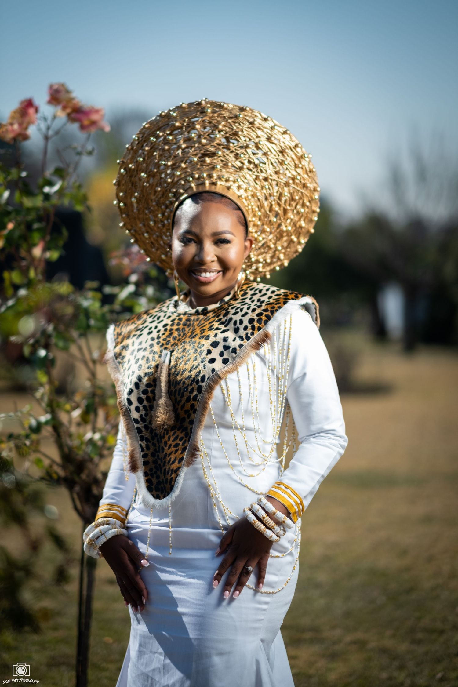 Traditional African-inspired fashion photoshoot featuring a gold headpiece and white dress in a garden setting.
