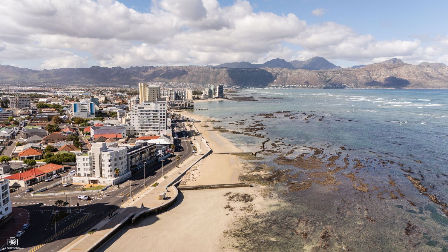 Coastal cityscape with buildings and harbor along rocky shoreline beneath mountain backdrop.