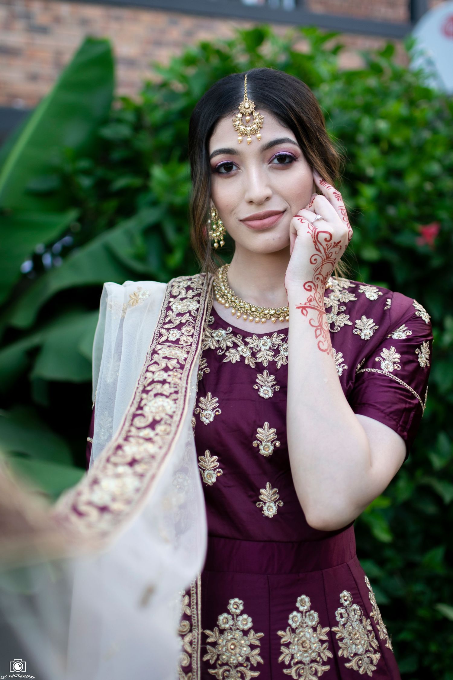 Traditional burgundy and gold outfit with jewelry photographed against garden backdrop.