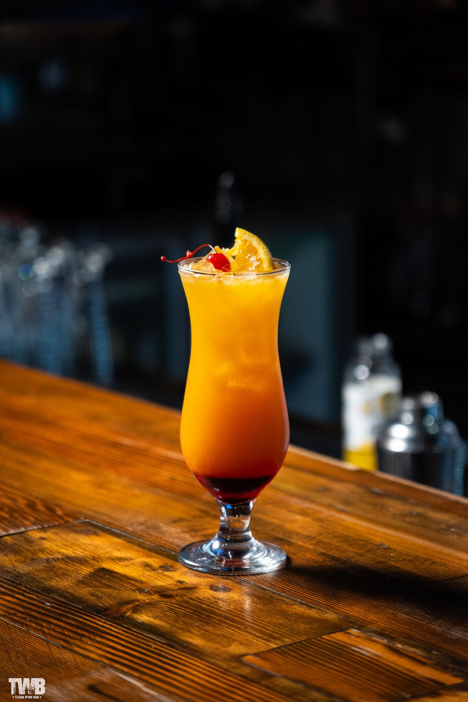 Tropical orange cocktail garnished with fruit in a hurricane glass on wooden bar top.