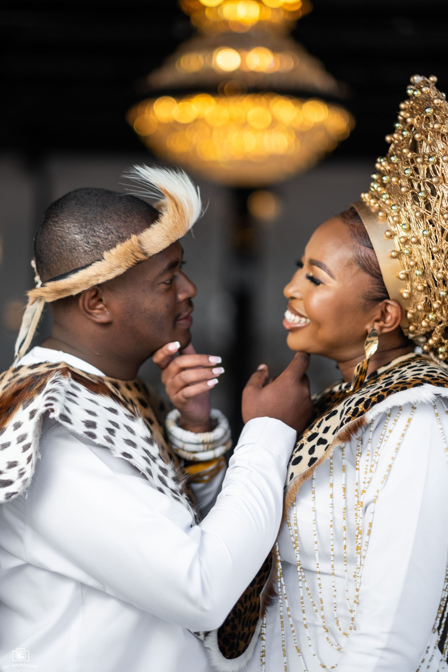 Couple in white and gold attire sharing romantic moment under chandelier lighting.