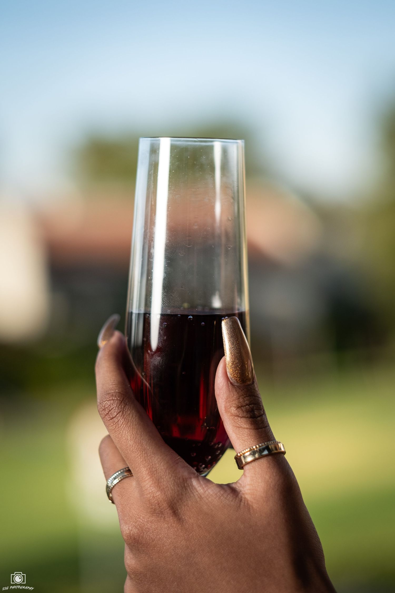 Hand holding a glass of red wine against a blurred outdoor background.