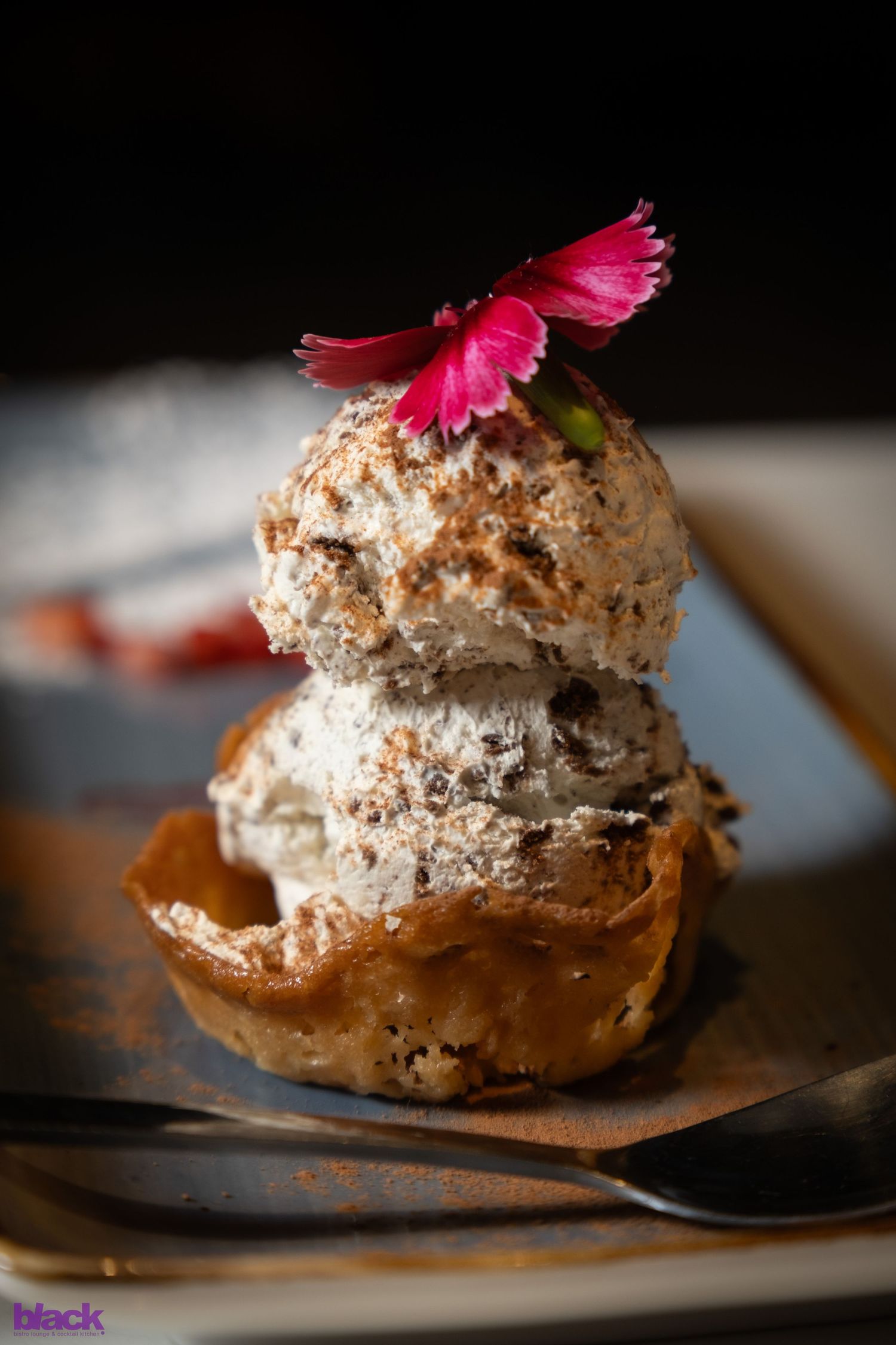 Gourmet dessert pastry topped with a pink flower on a black plate.