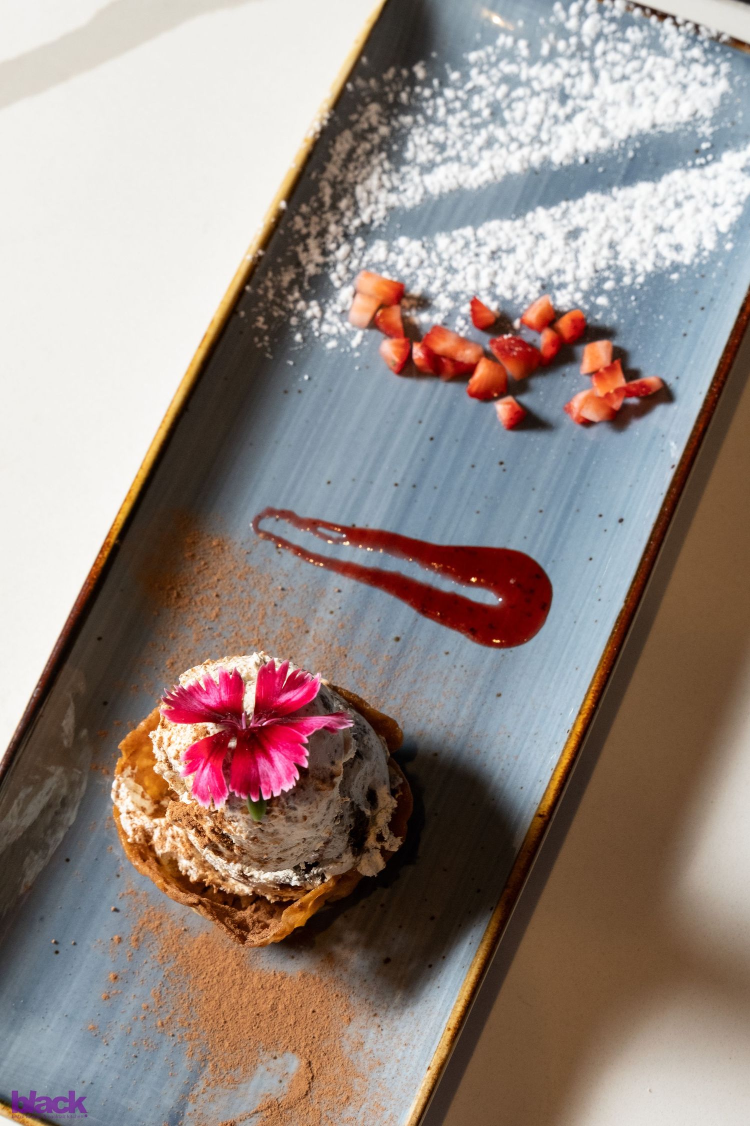Plated dessert with pink flower garnish and red sauce drizzle on grey slate.