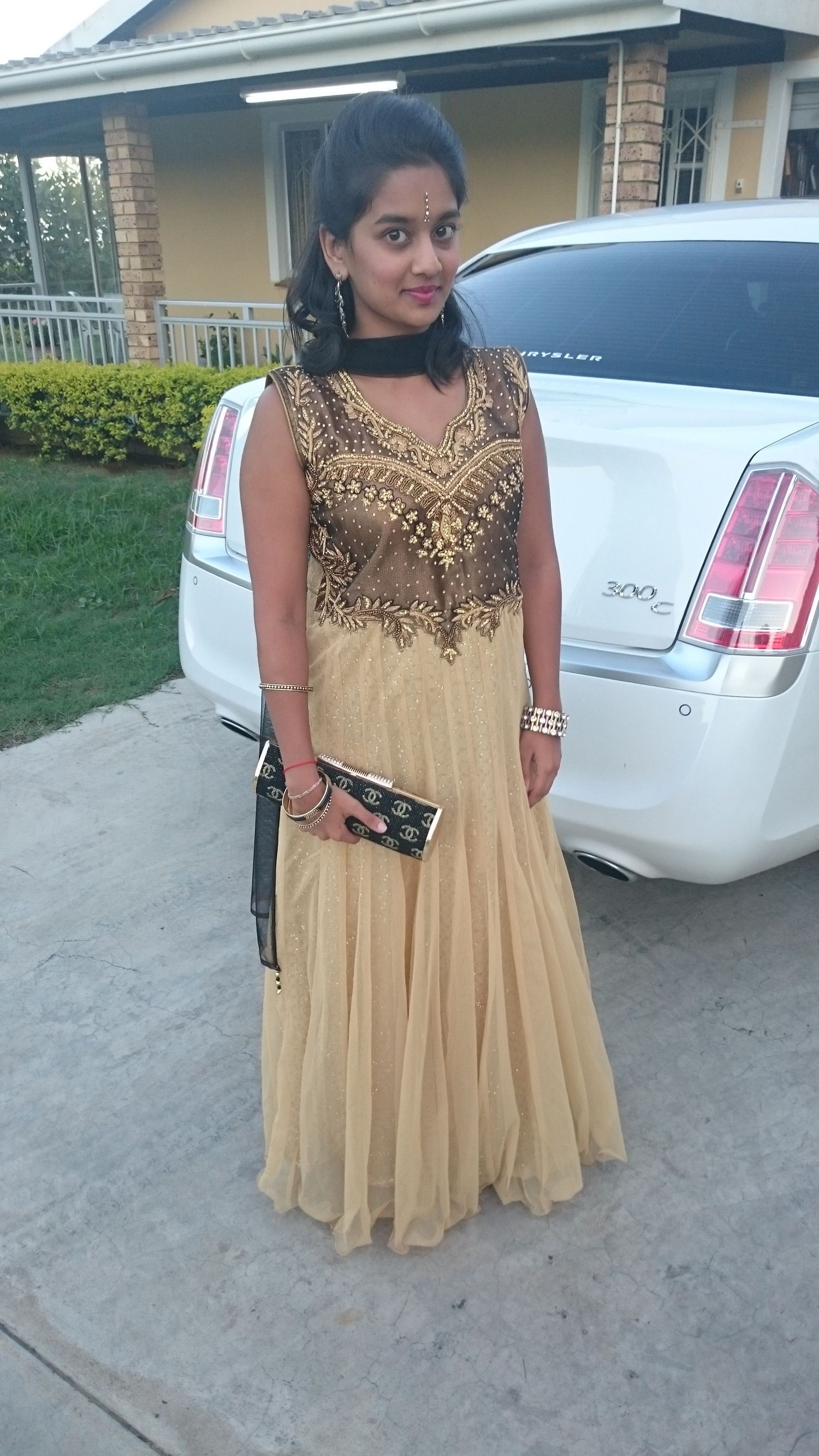 A person wearing a beige and gold embellished sleeveless dress poses next to a white car in a driveway.