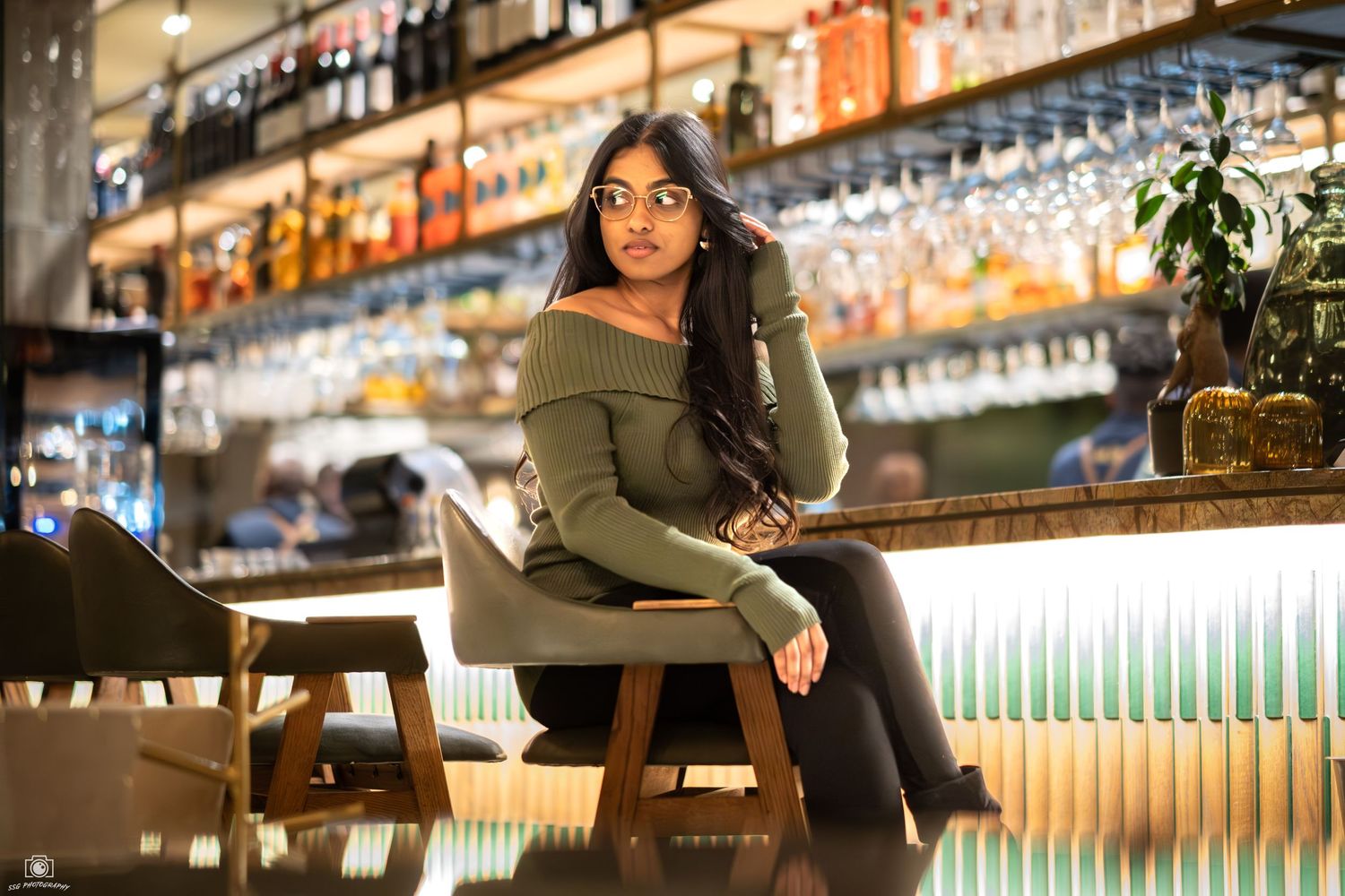A person in green sits at an elegant bar with hanging glasses and ambient lighting.