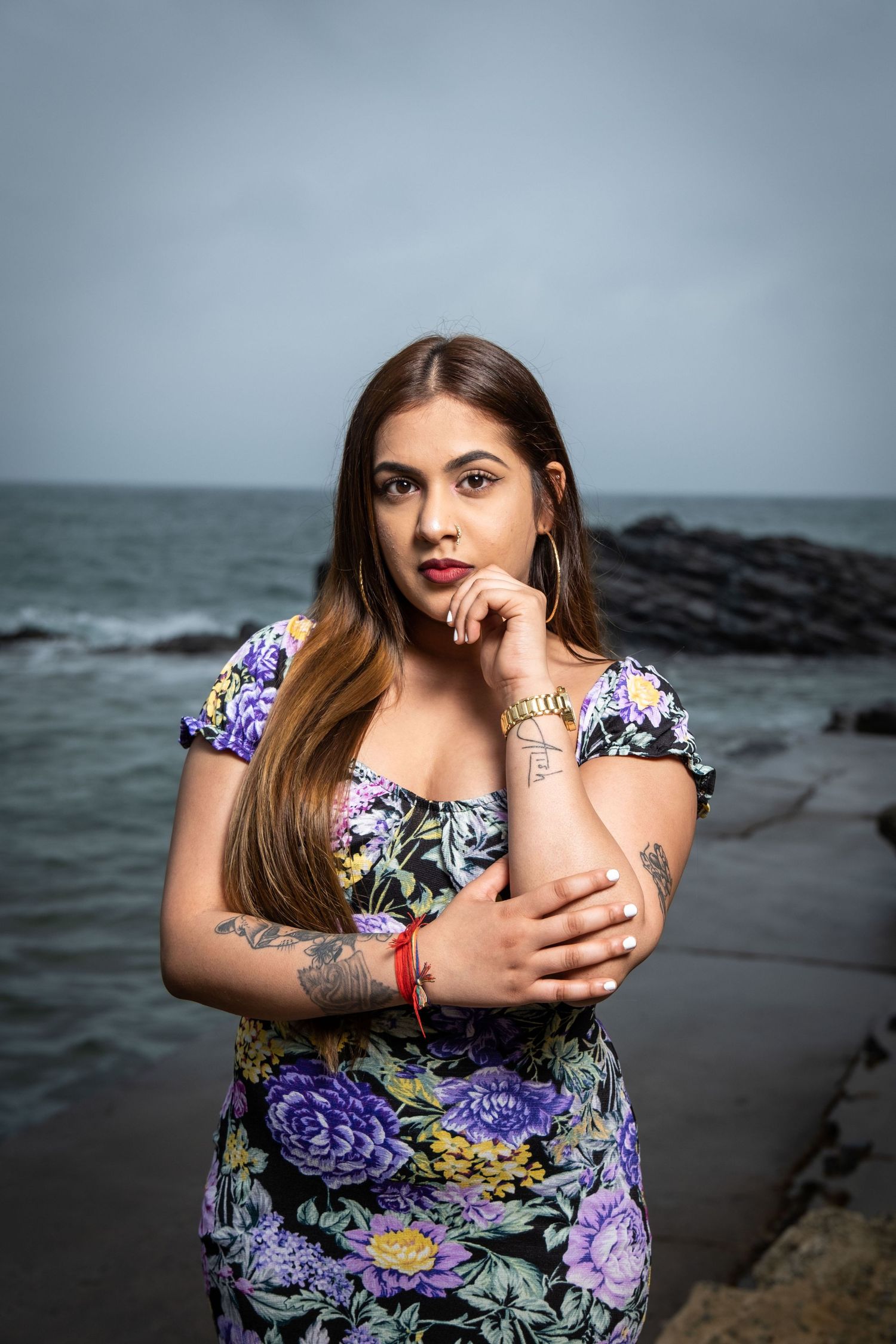 Woman in floral dress poses artistically by ocean with moody sky background.