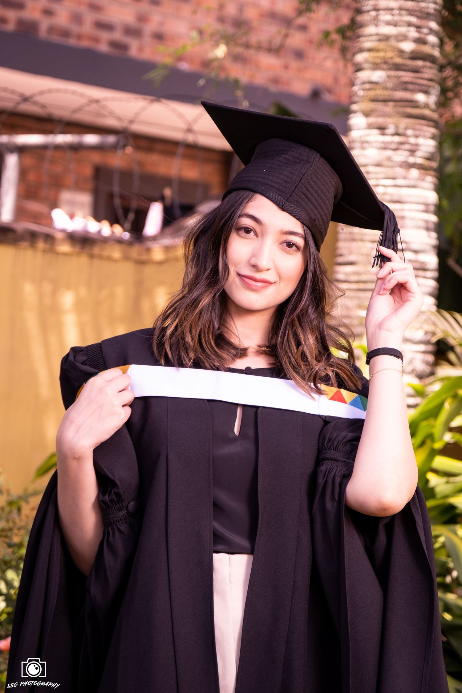 Graduate in cap and gown poses cheerfully with graduation attire against brick wall background.