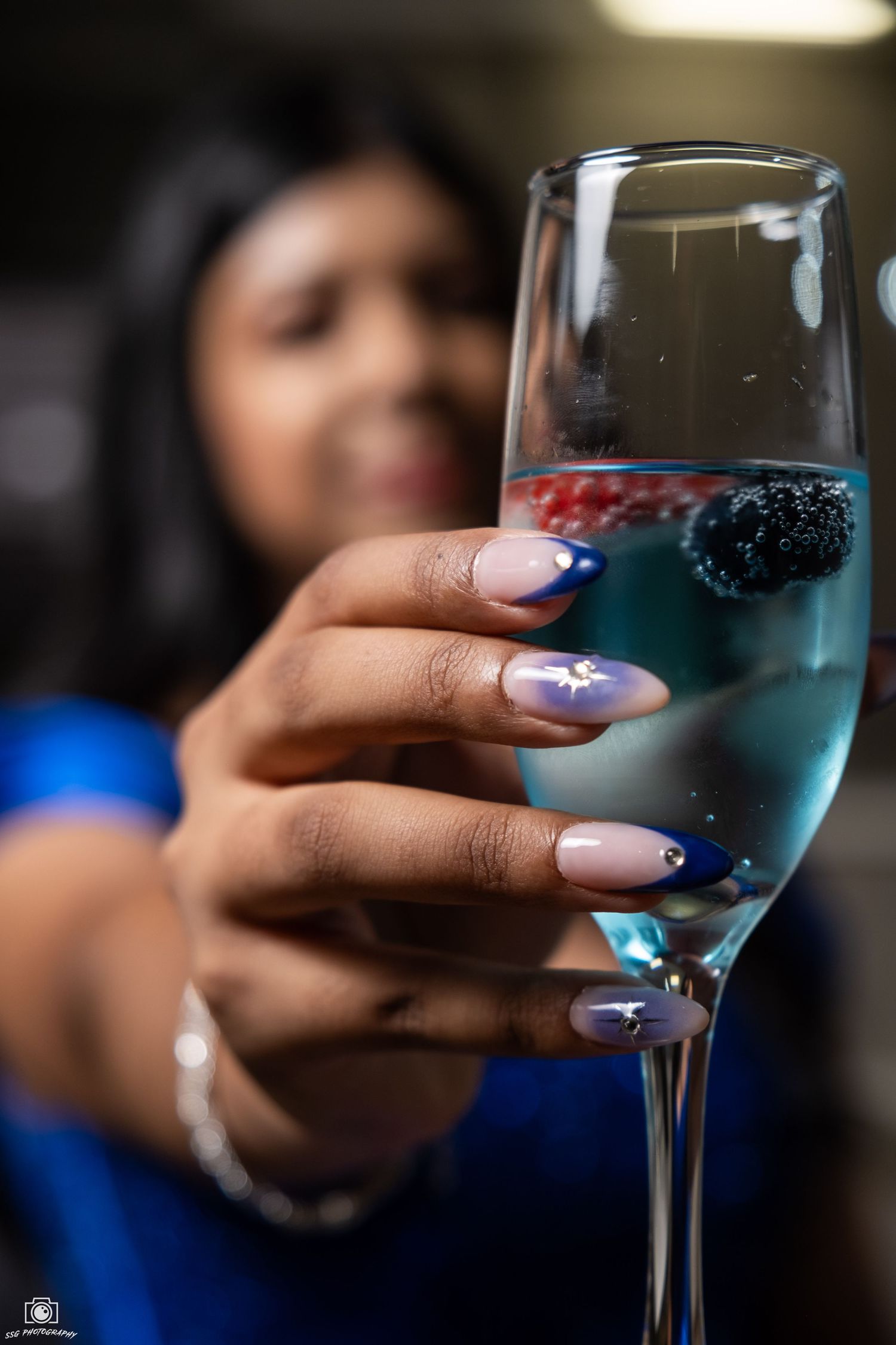 Manicured hand holds blue cocktail with berries in stemmed glass against blurred background.