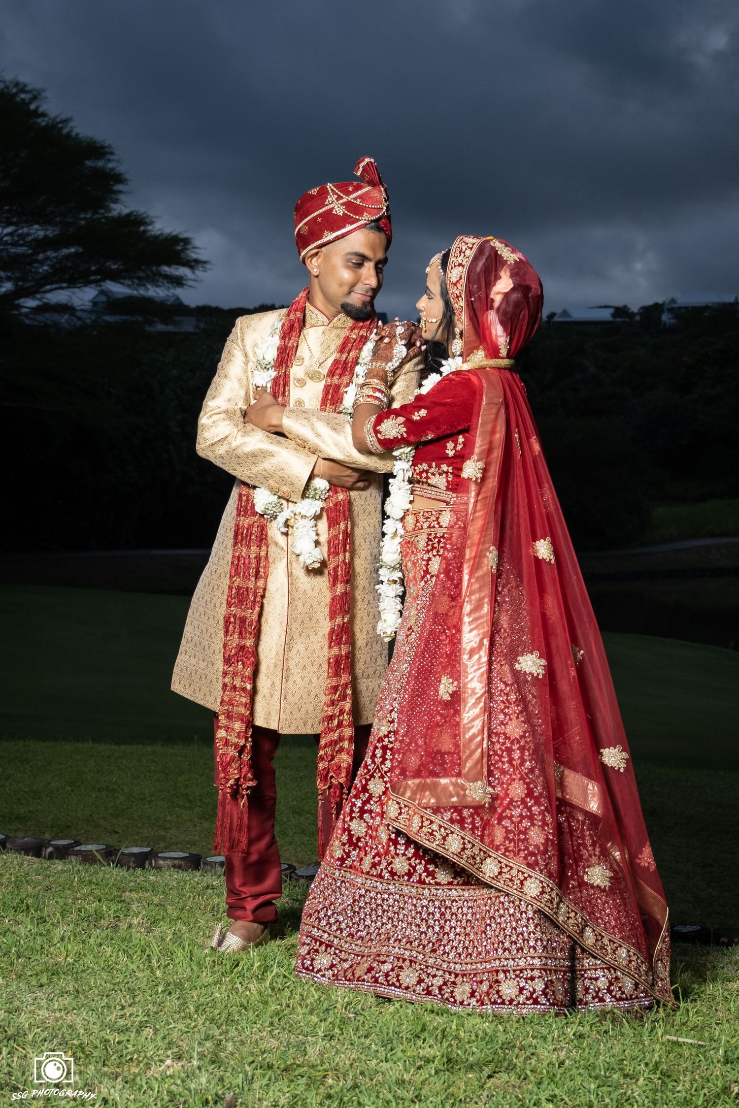Couple in traditional Indian wedding attire posing on grass with dramatic sky.