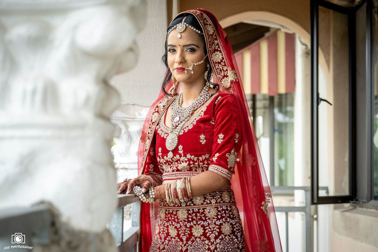 Bride in traditional red Indian wedding attire near window with soft lighting.