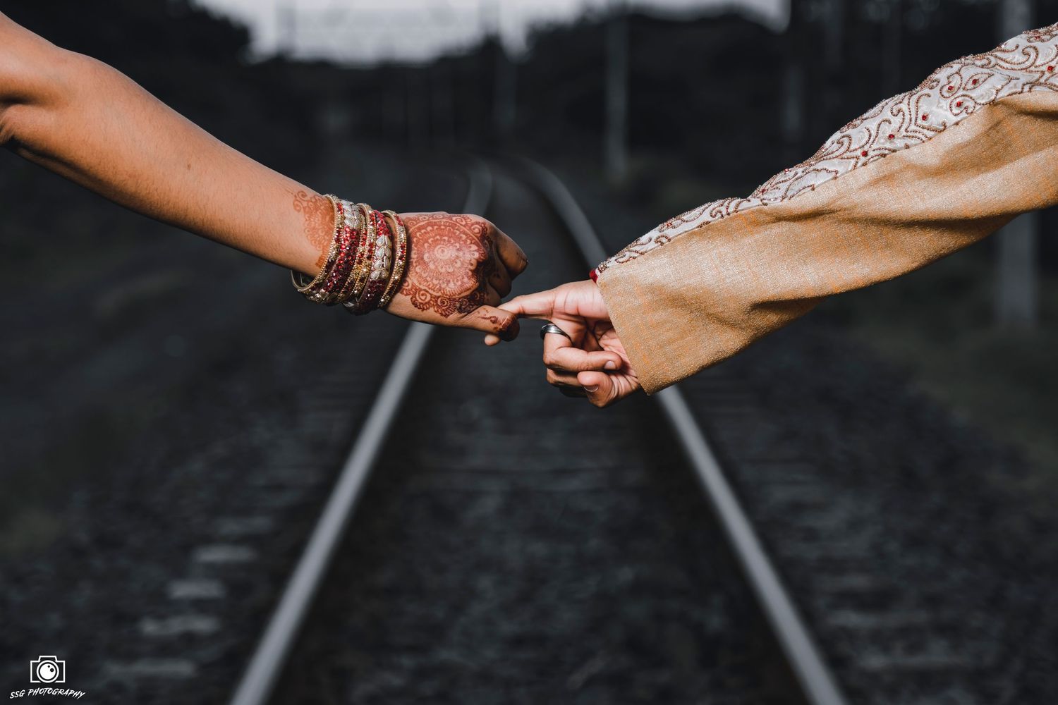 Two hands adorned with traditional Indian wedding jewelry reach out to hold each other against a backdrop of railway tracks.