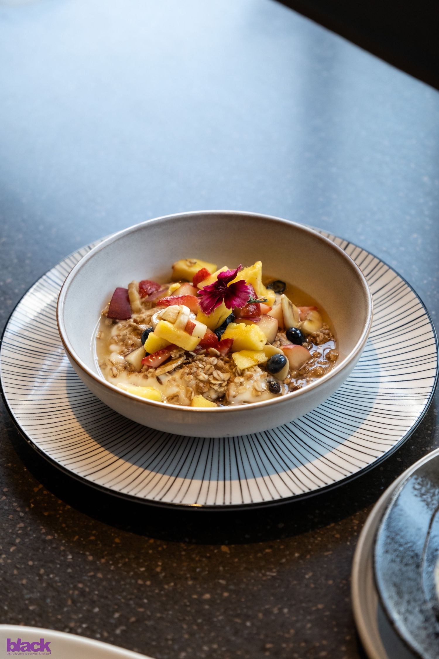 Elegant breakfast bowl with tropical fruits and granola served on white striped plate in natural lighting.