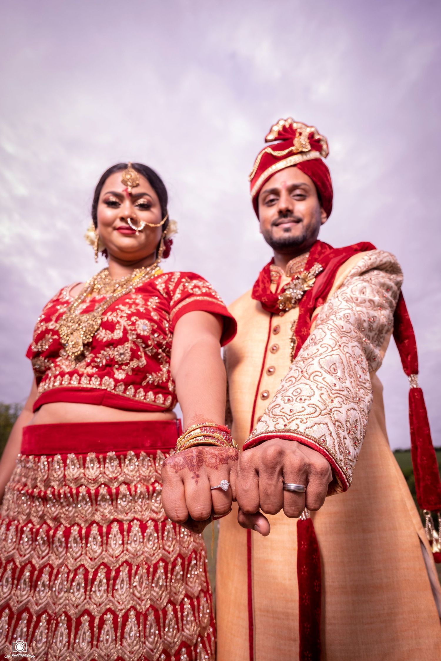 Traditional Indian wedding couple in red and cream attire show off their rings against a purple sky.