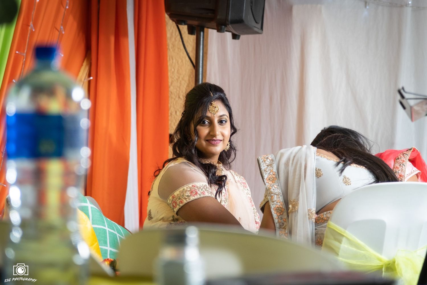 Bride in traditional Indian attire with henna decorated hands poses against colorful draped backdrop.
