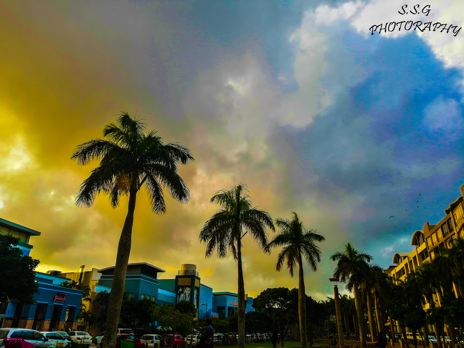 Dramatic sunset sky over palm trees and buildings silhouetted against vibrant yellow and blue clouds.