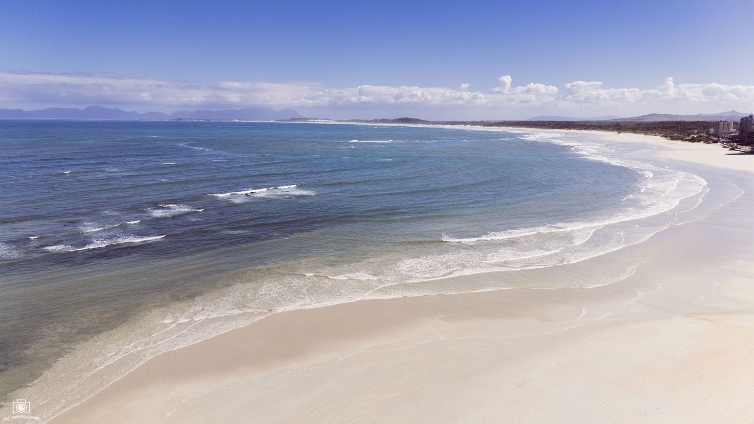 Sweeping aerial view of curved white sandy beach with rolling waves and blue ocean.