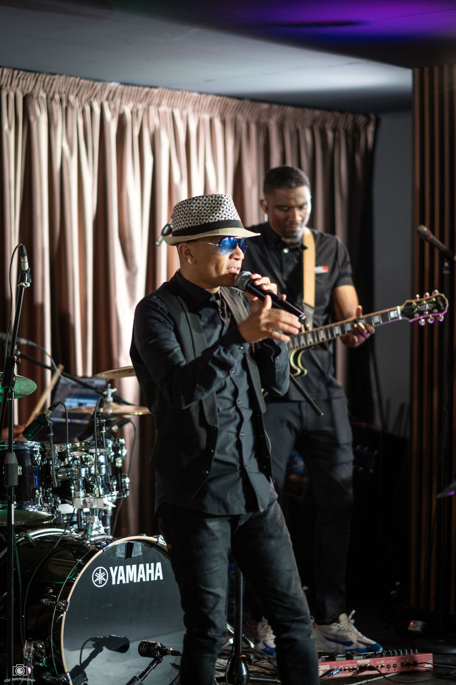 Performer in black attire and fedora sings on stage with a Yamaha drum kit and band member visible in background.