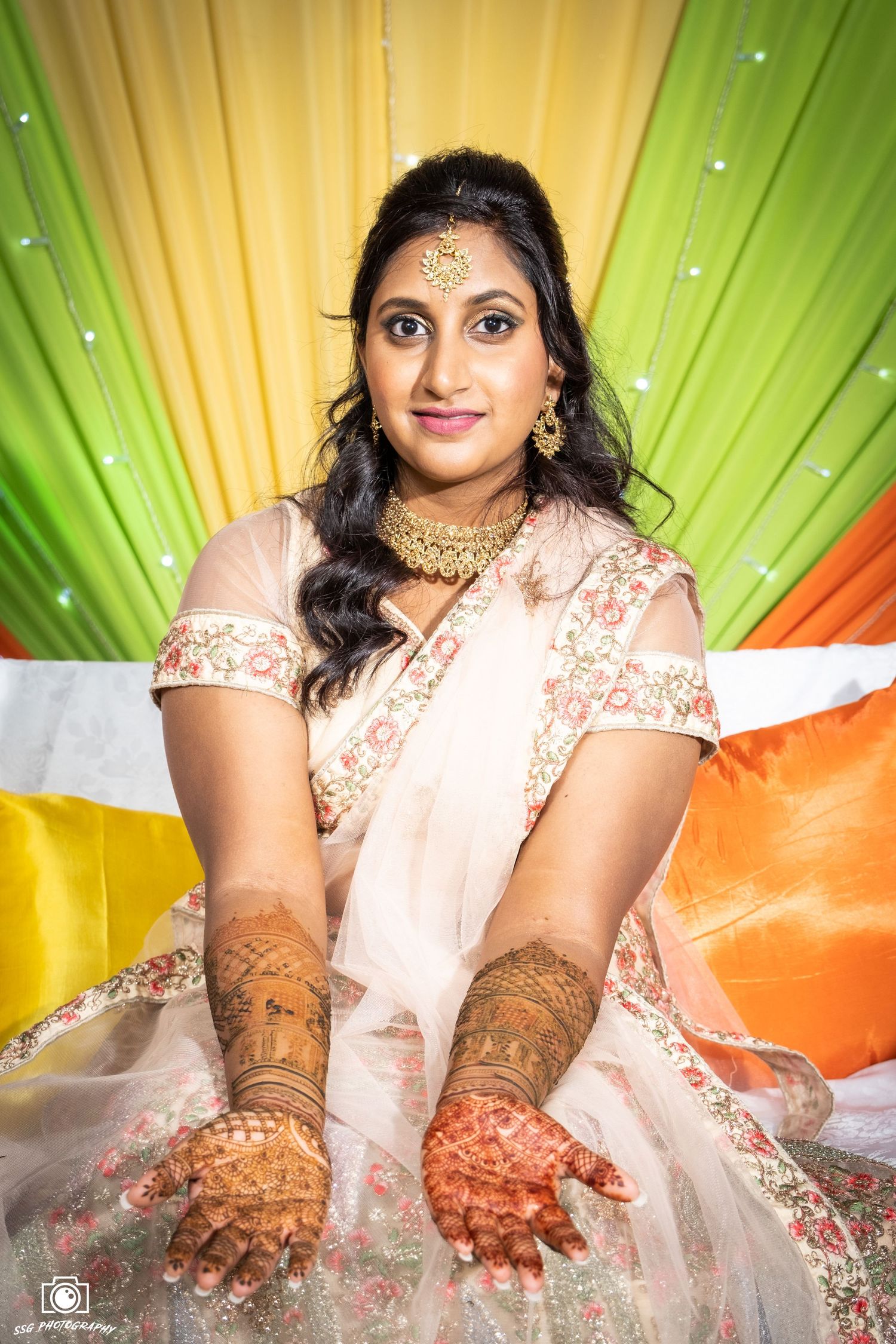 Bride in embroidered outfit displays mehndi designs against orange and green backdrop.