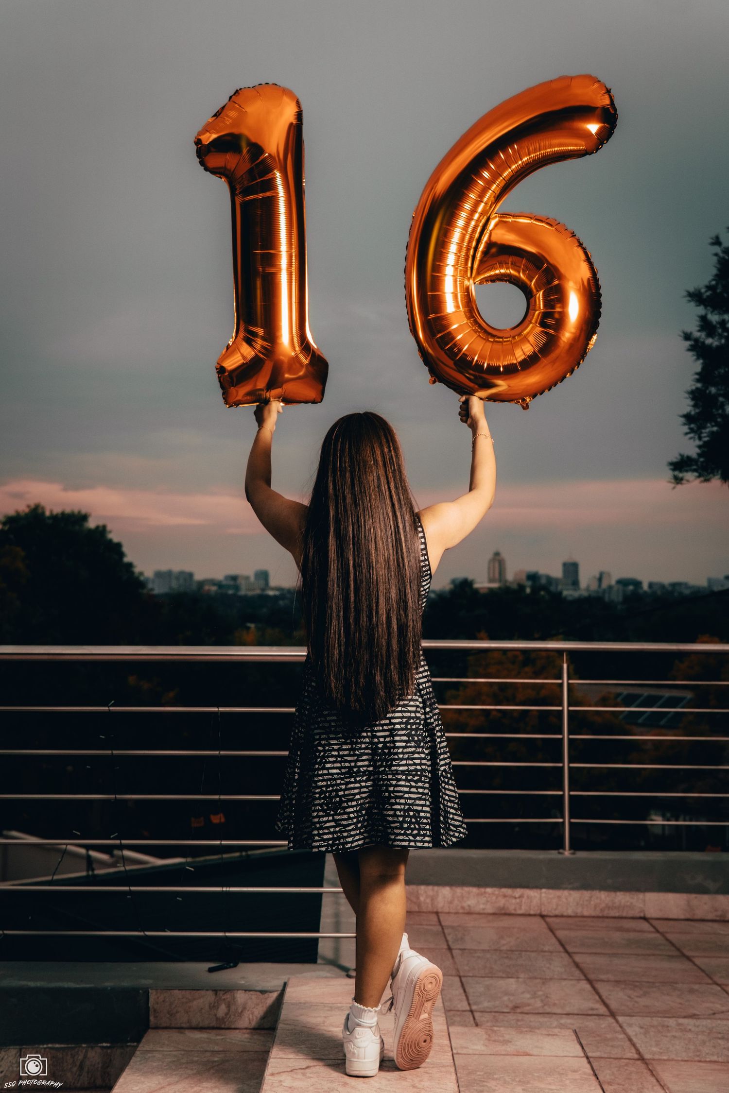 Figure holding gold number 16 balloons against sunset city skyline.
