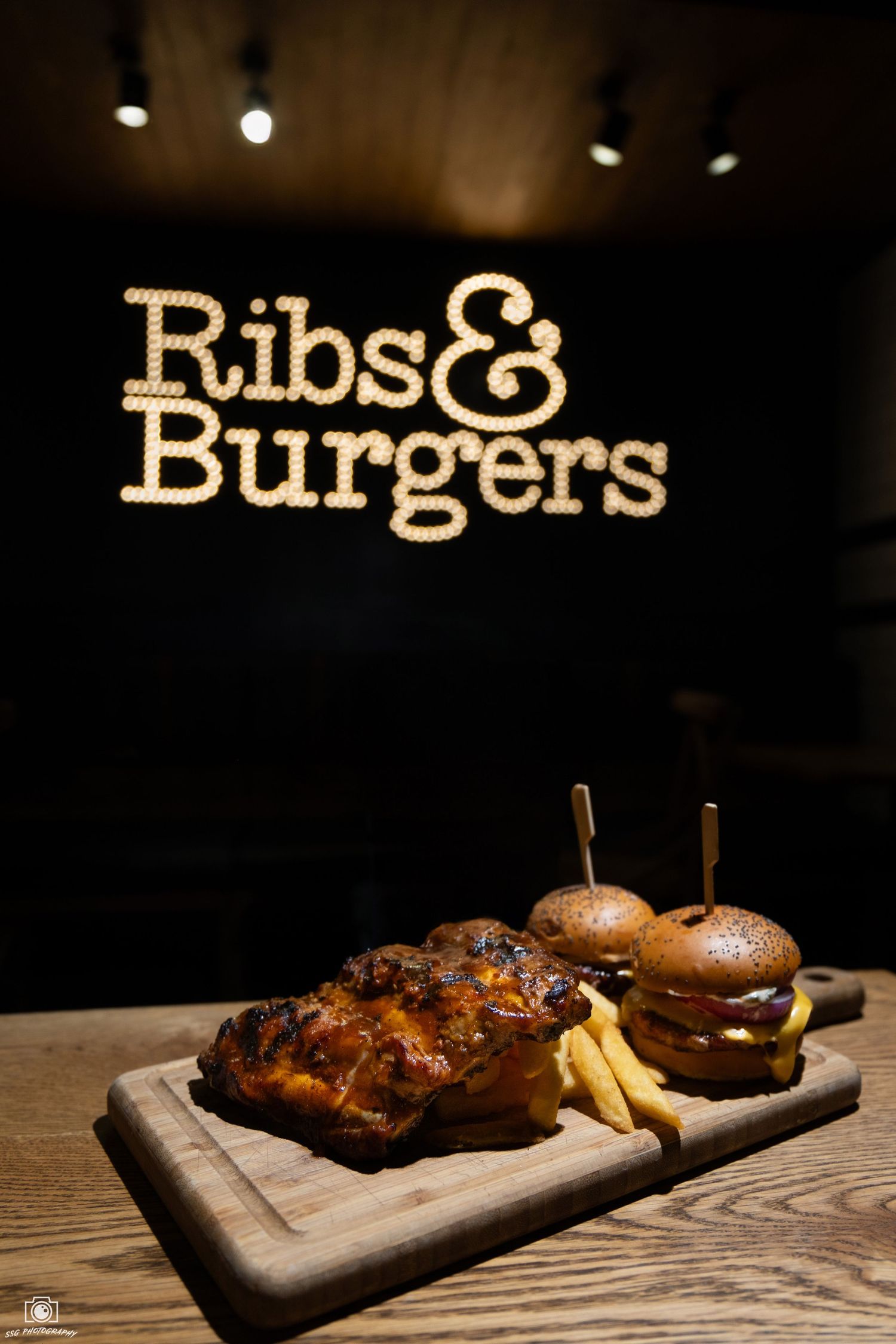 Wooden serving board with burgers and fries under illuminated restaurant signage.