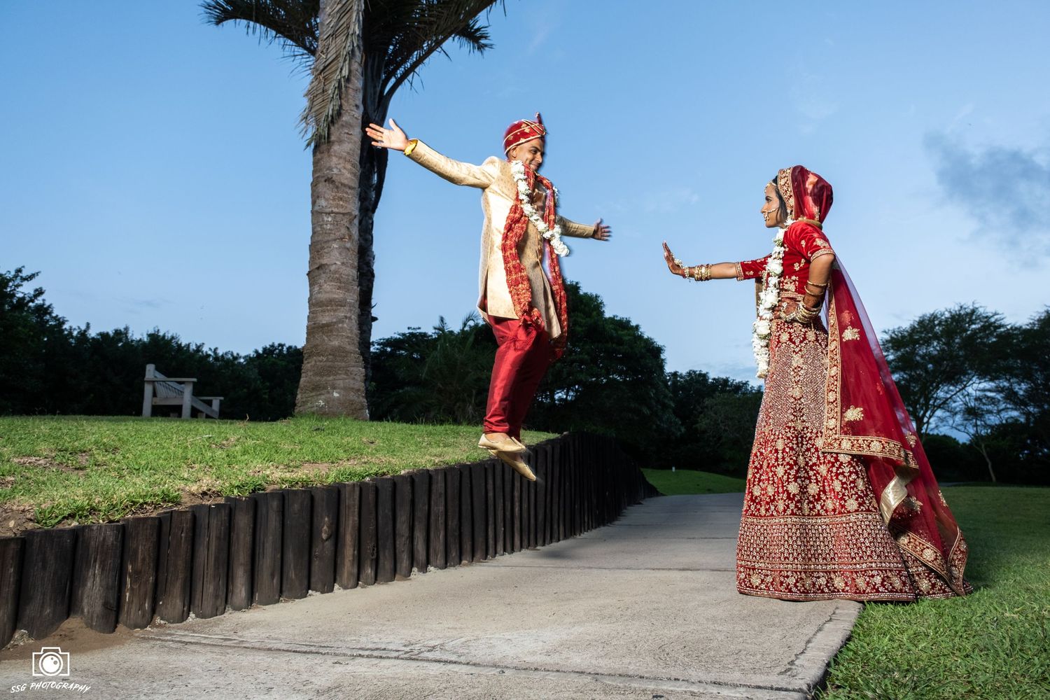 Wedding photo session on a palm-lined pathway with traditional red and gold attire.