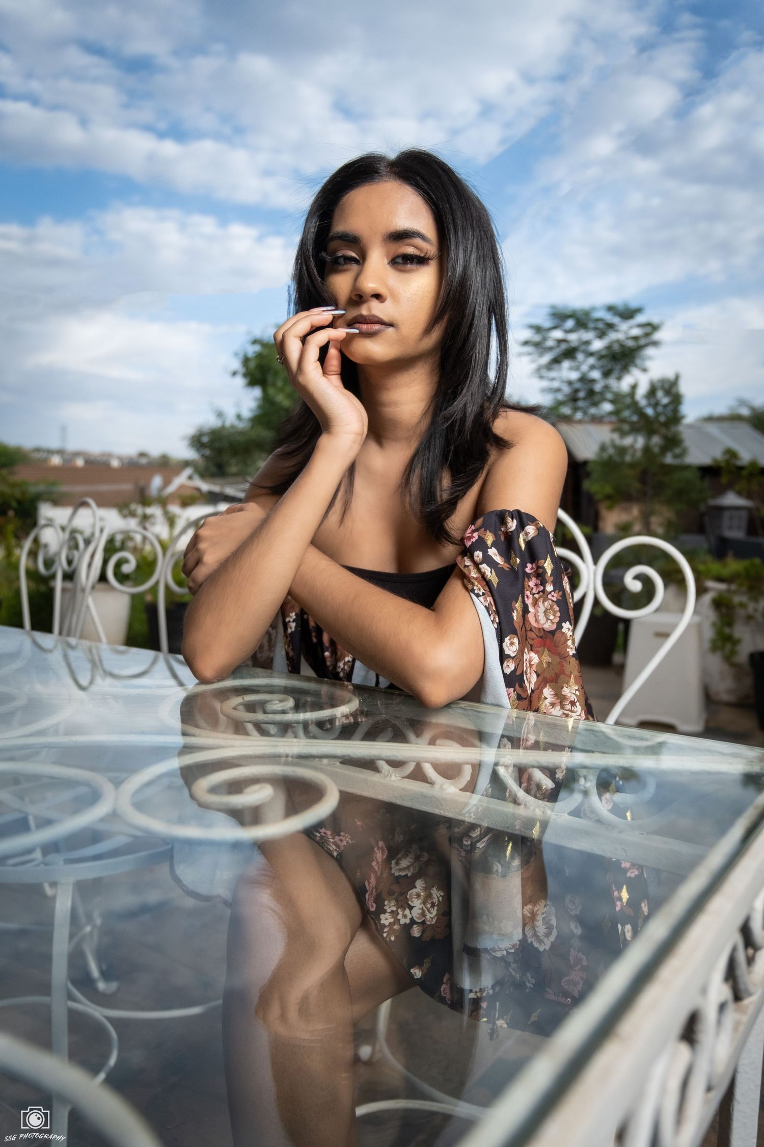 Fashion model in black lace dress poses against ornate white railing outdoors with blue sky background.