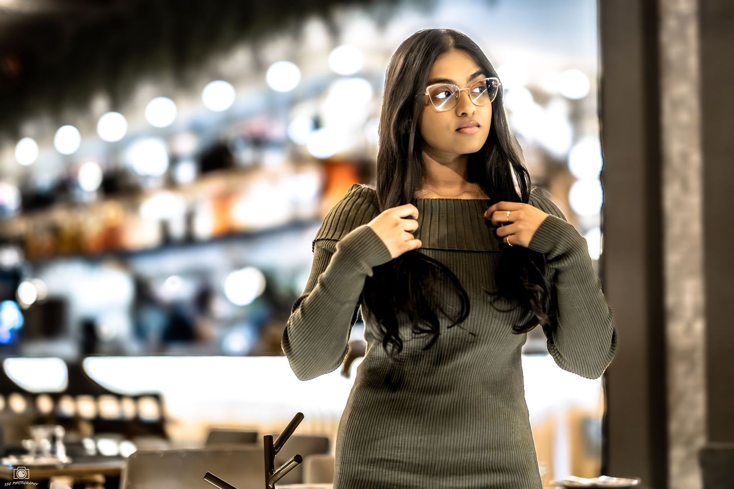 A person in a green sweater sits at a bar counter with glowing lights and glassware in the background.