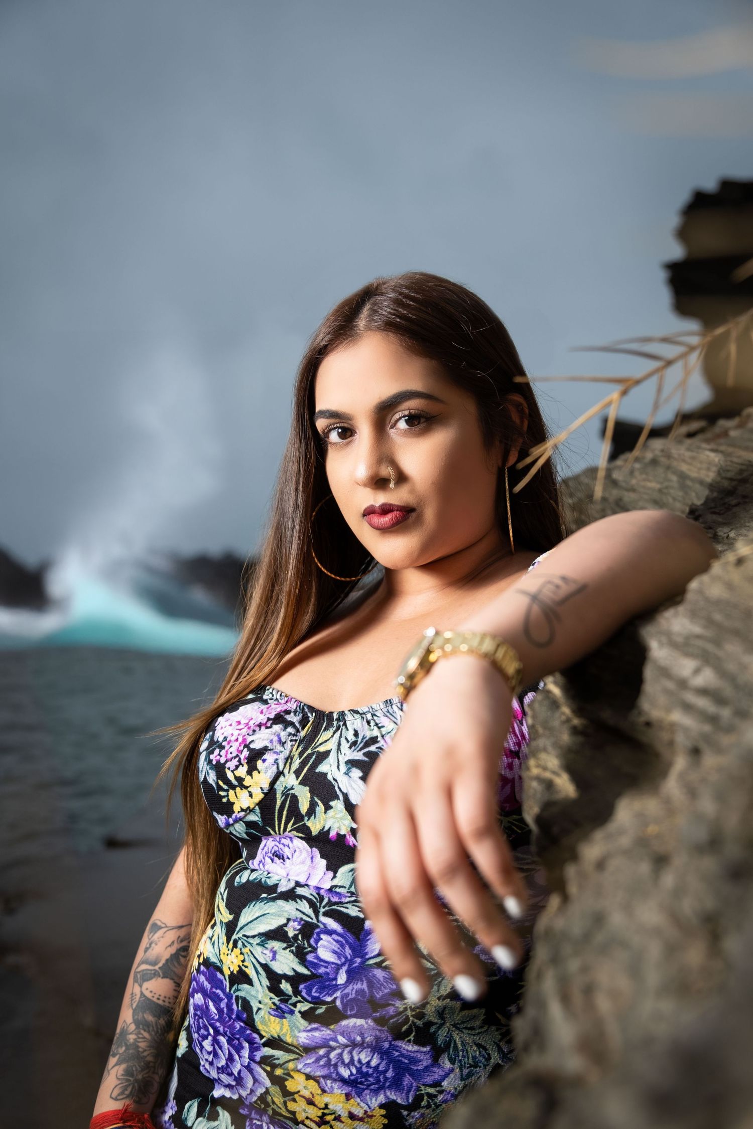 Model in purple floral dress poses dramatically against rocky coastal backdrop.