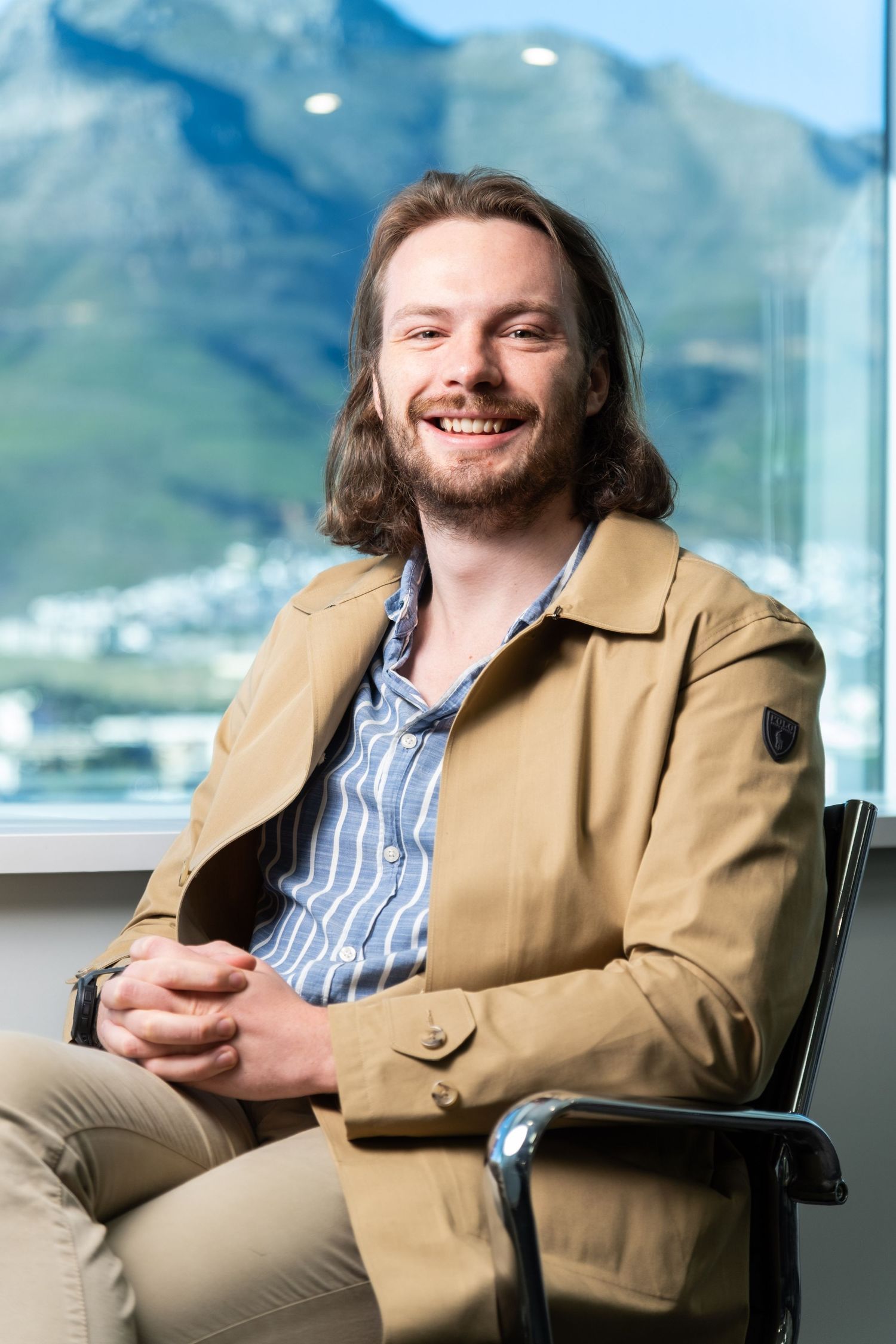 Professional in tan jacket seated casually in modern office with mountain view.
