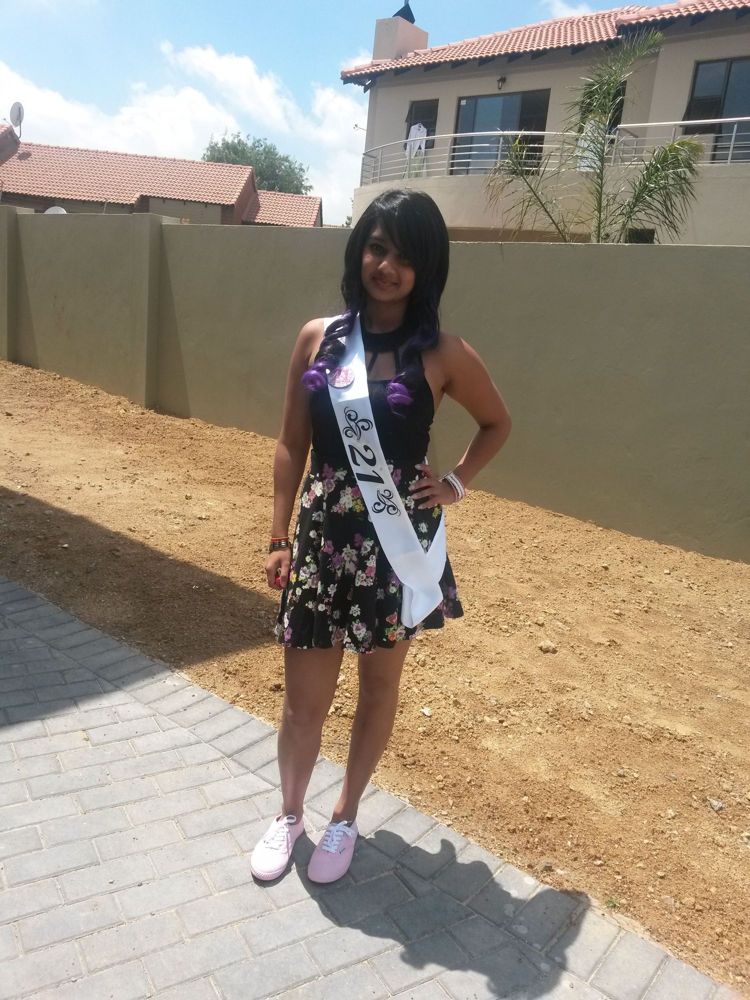 A person in a floral dress and sash poses outdoors against a wall on a sunny day.